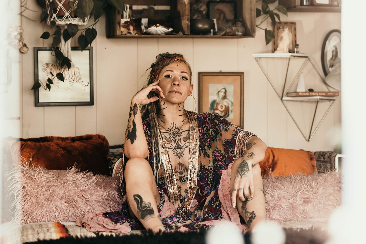 Heavily tattooed woman sits relaxed on couch in eclectic bohemian decorated room.