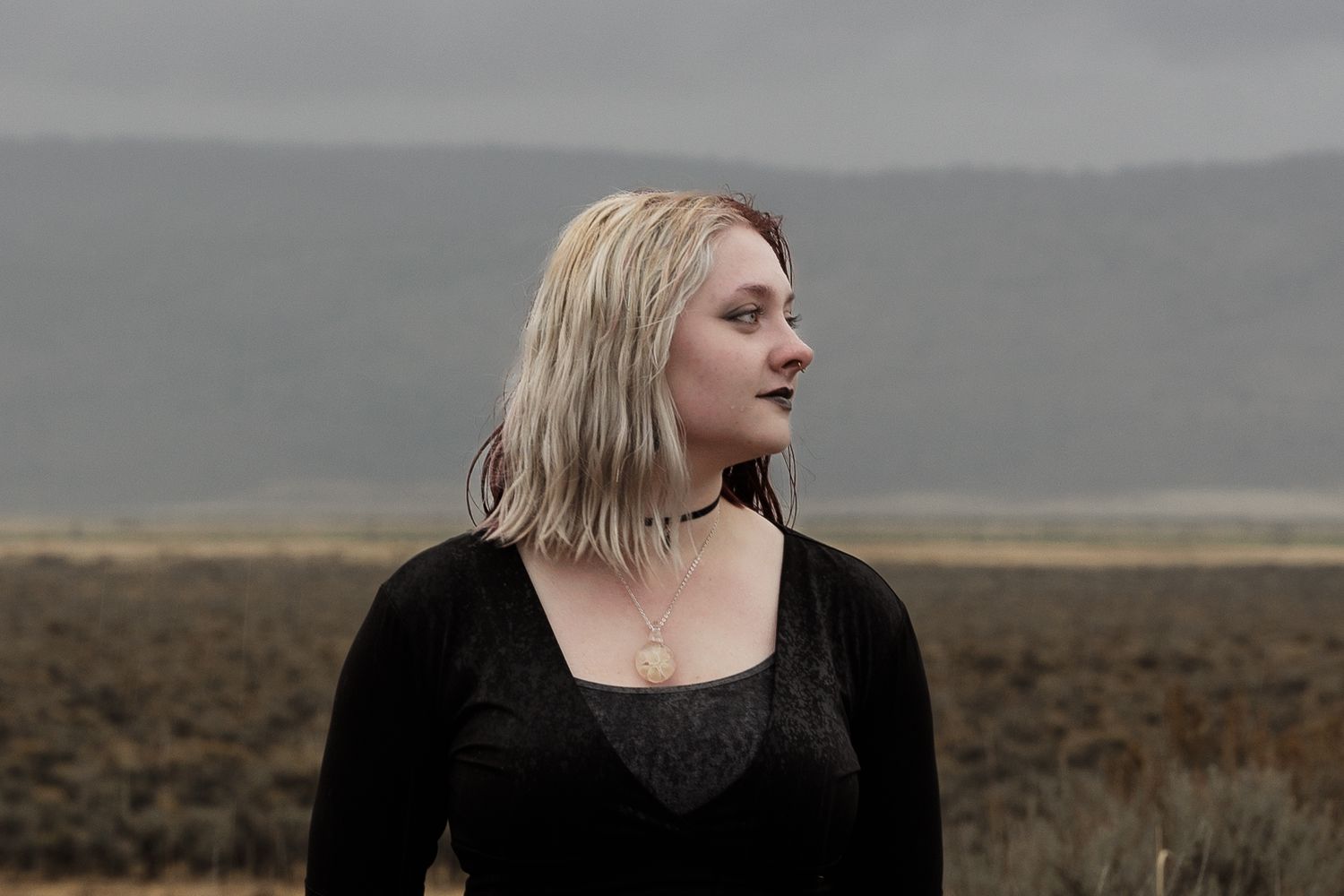 Young blonde woman in black outfit standing in an open moorland landscape under a grey cloudy sky.