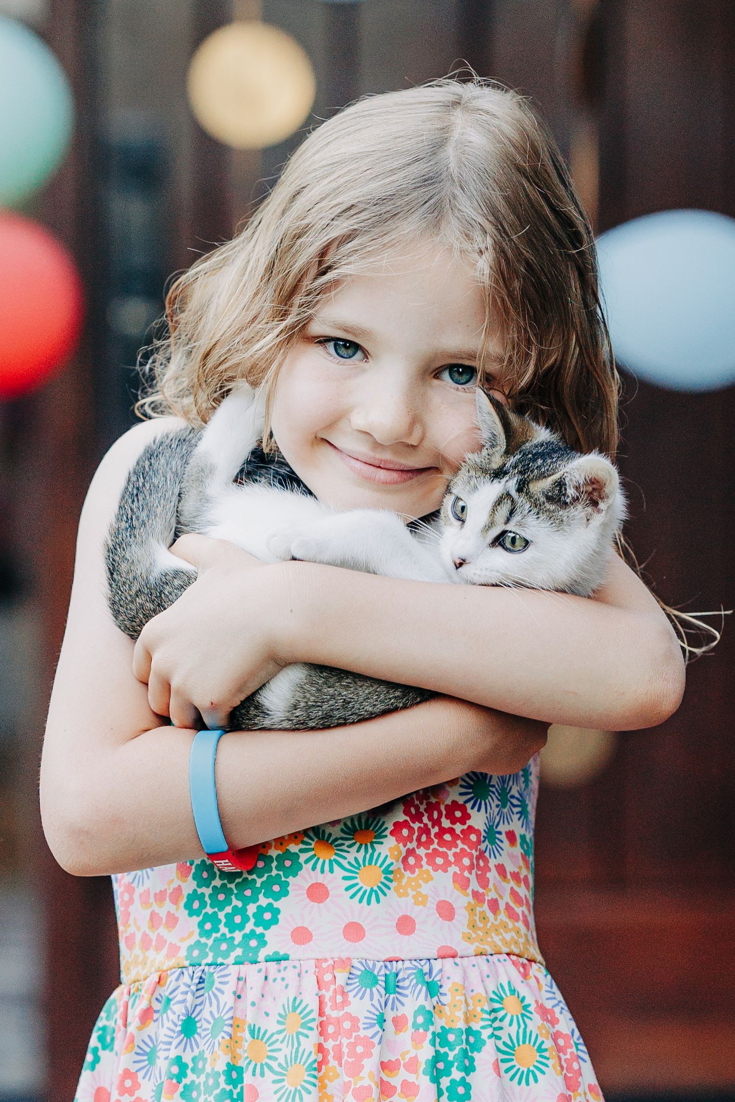 Sweet young girl in floral dress smiles while tenderly hugging adorable gray and white kitten at party.
