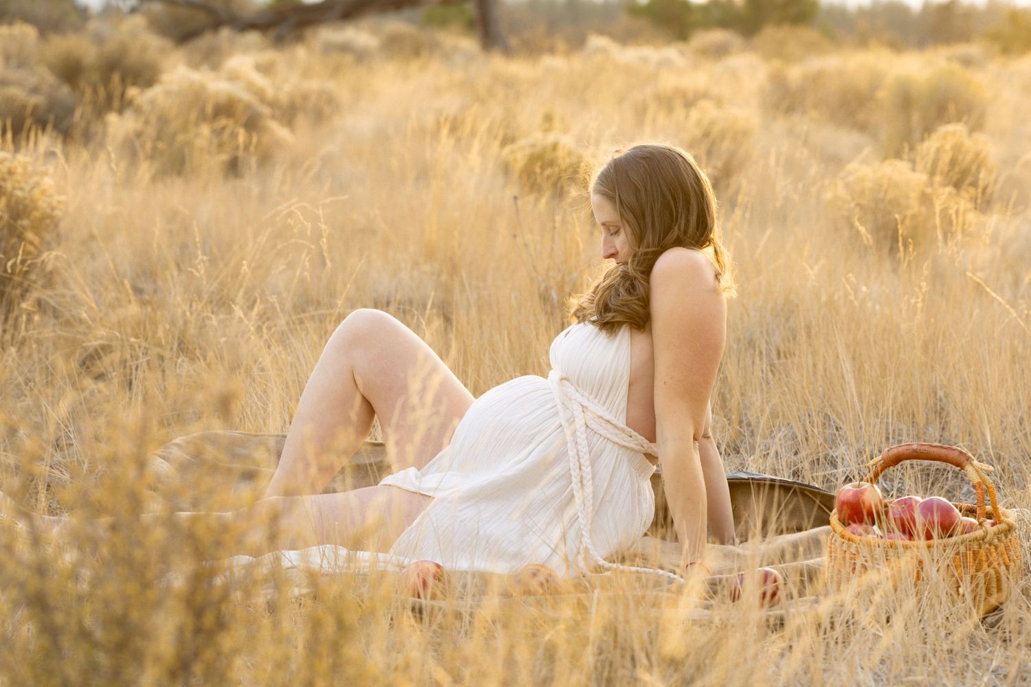 Pregnant woman in white dress relaxing in golden dry grass field during sunset maternity session.