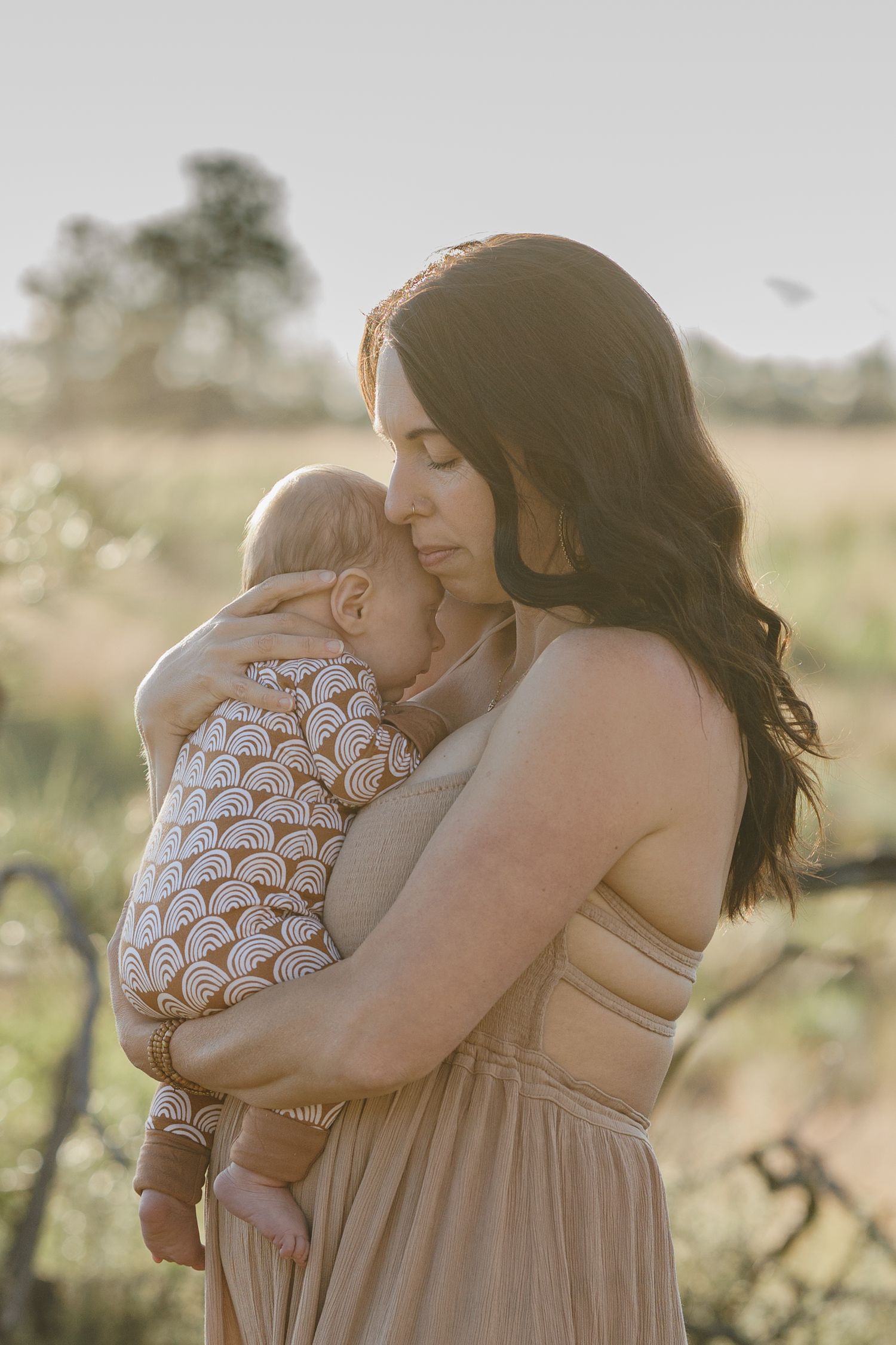 Mother in beige strapless dress tenderly embracing newborn baby outdoors in warm golden sunlight.