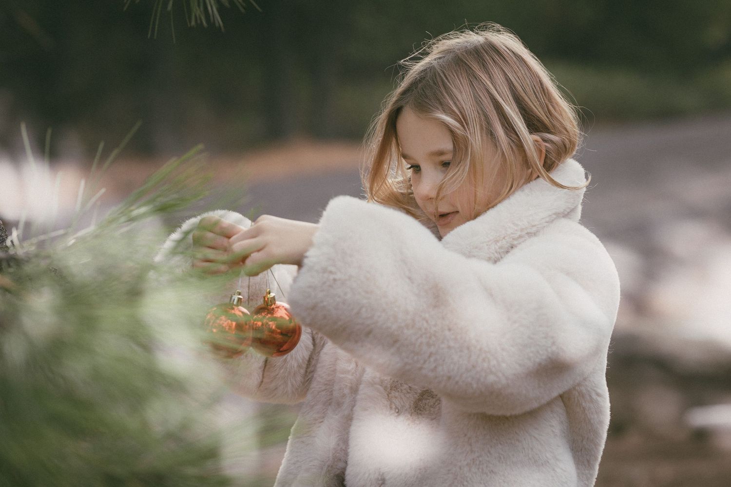 Young blonde girl in white fluffy coat examining Christmas ornament on pine tree branch outdoors.