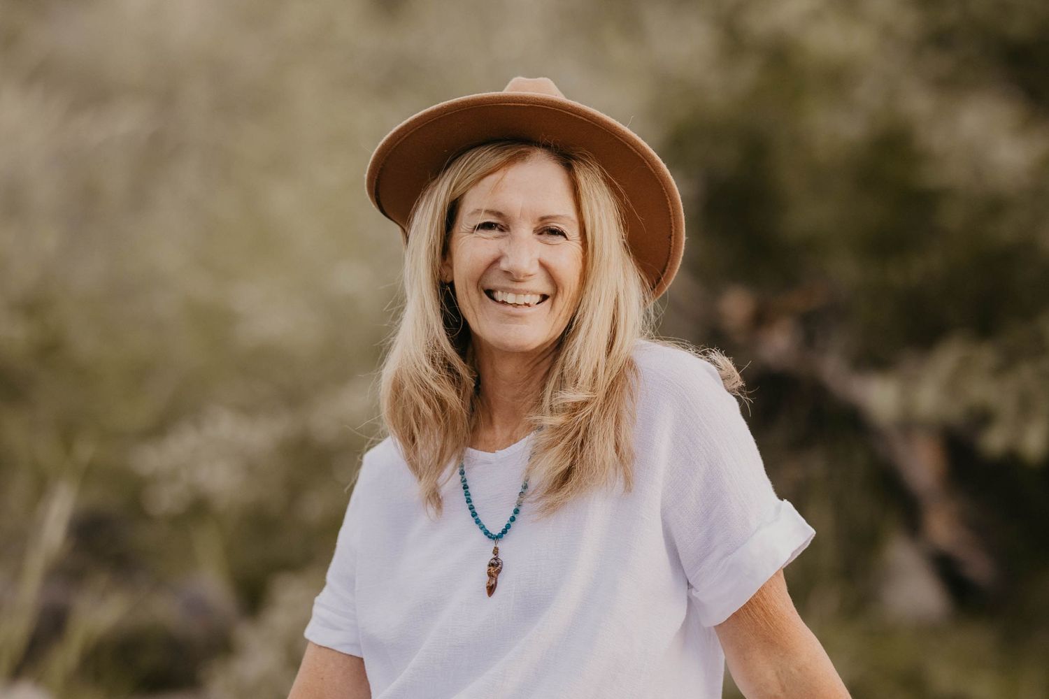 Smiling blonde woman in white tee and brown hat outdoors.