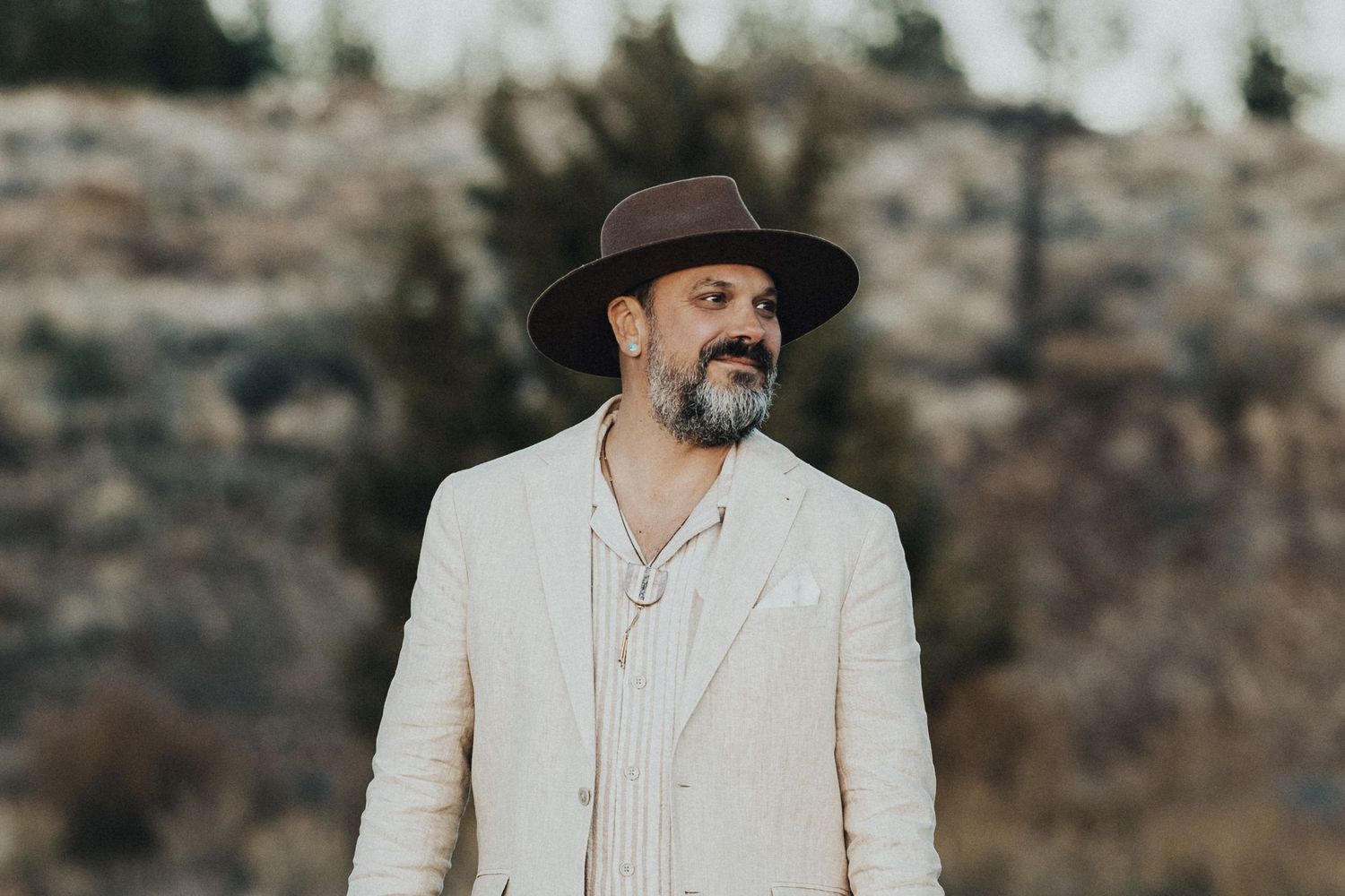 Bearded man in wide-brim hat and linen suit gazing into distance in outdoor rocky landscape.