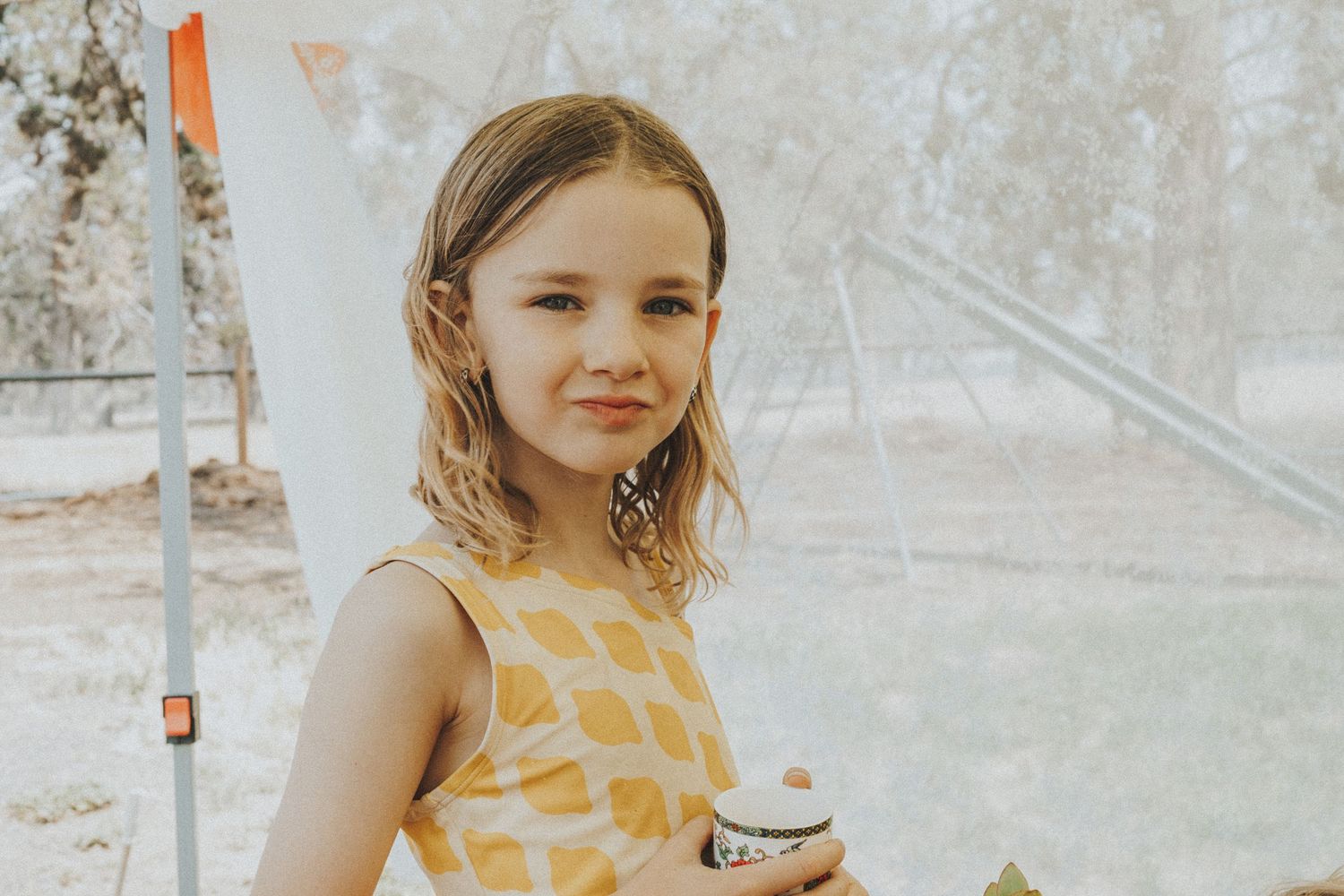 Young girl in yellow patterned dress holding cup outdoors near tent.