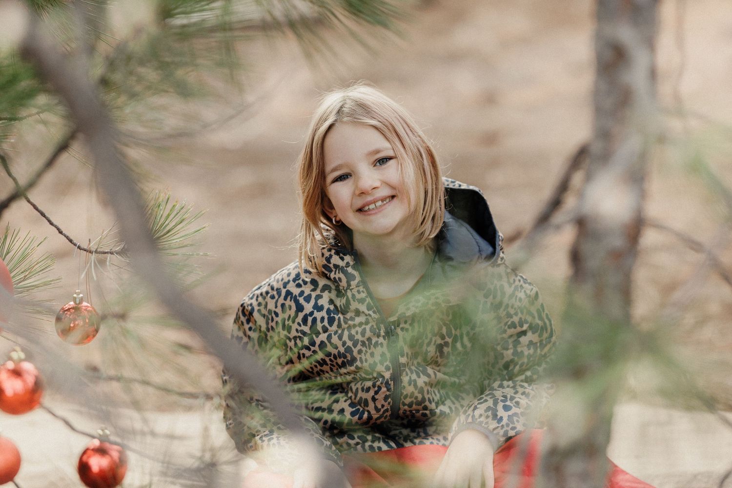 Smiling blonde girl in leopard print jacket peeking through pine branches decorated with red ornaments.