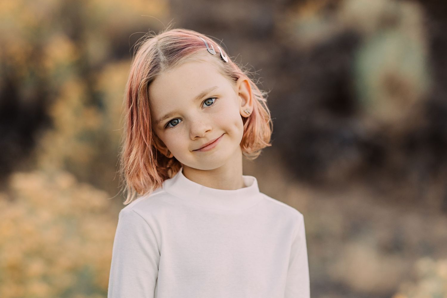 Smiling young girl with pink-tipped hair in white turtleneck posing outdoors in warm light.