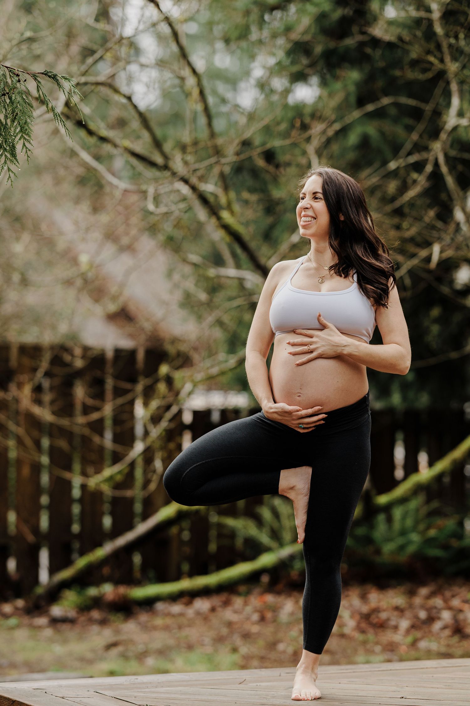 Smiling pregnant woman doing a tree yoga pose outdoors while cradling her baby bump.