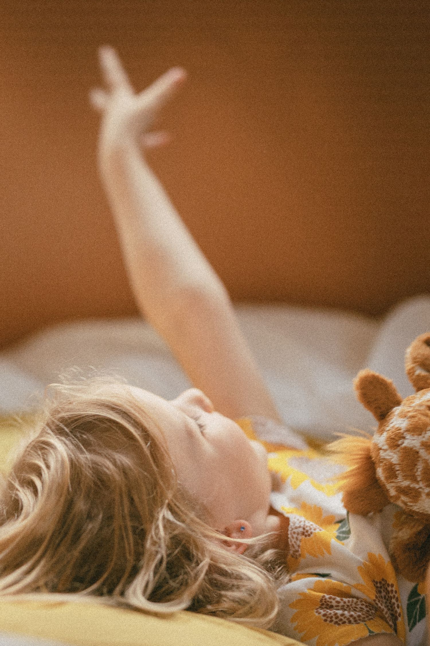 Young blonde girl lying on bed reaching her arm up toward the ceiling next to a stuffed giraffe toy.
