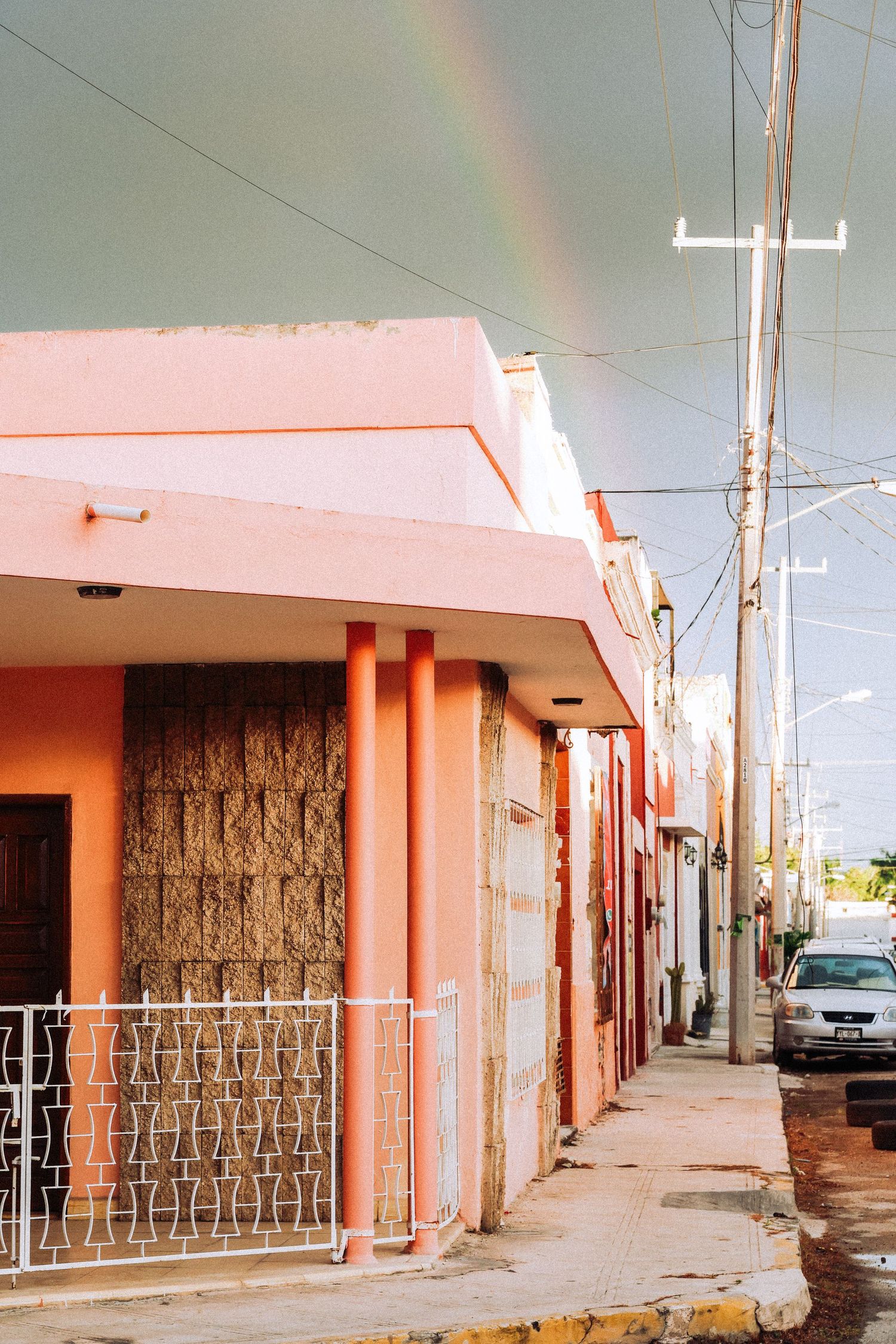 Pink and orange building on a narrow sidewalk street with a rainbow visible in the cloudy sky above.