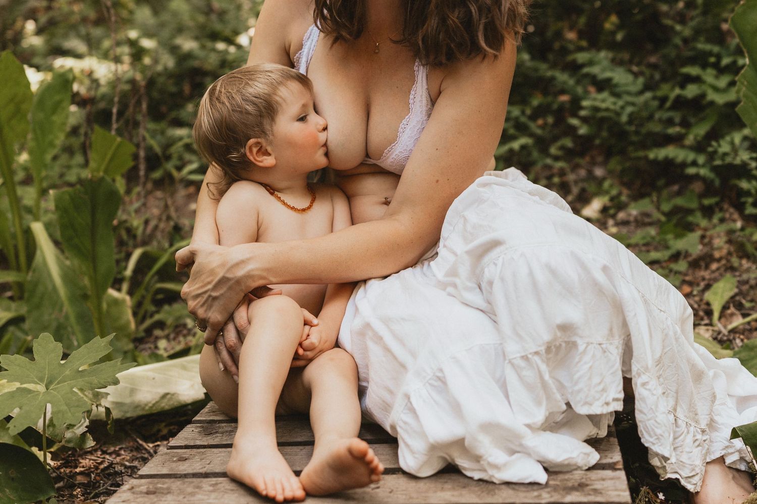 Mother breastfeeding toddler outdoors in lush garden setting, wearing white skirt, natural parenting lifestyle photography.