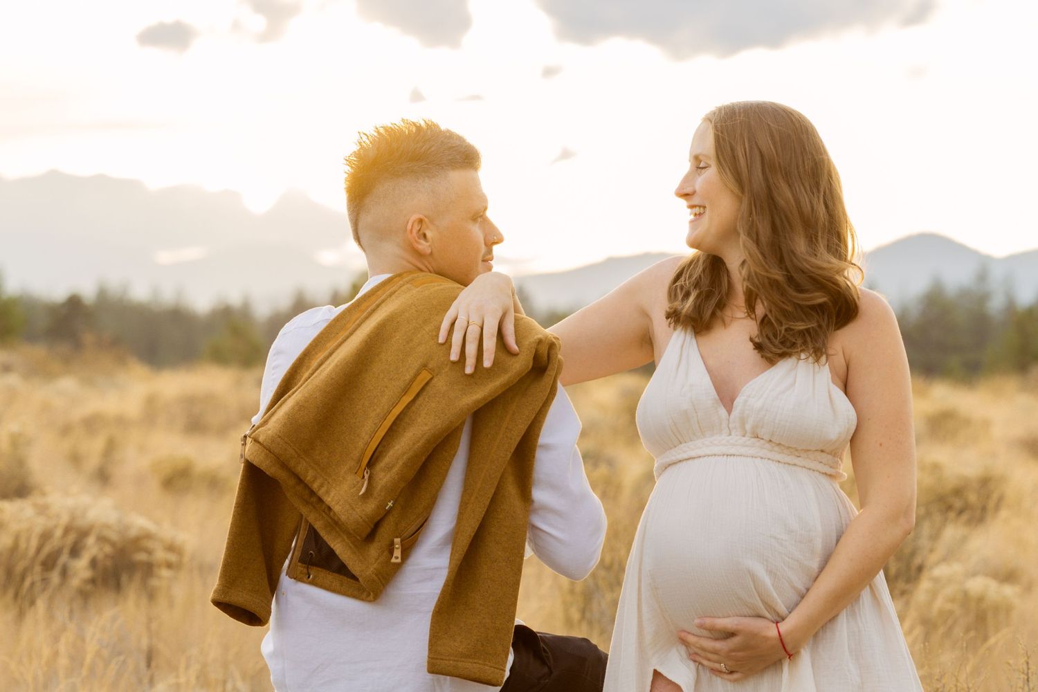 Smiling pregnant woman in white dress with partner in golden field at sunset during maternity shoot.