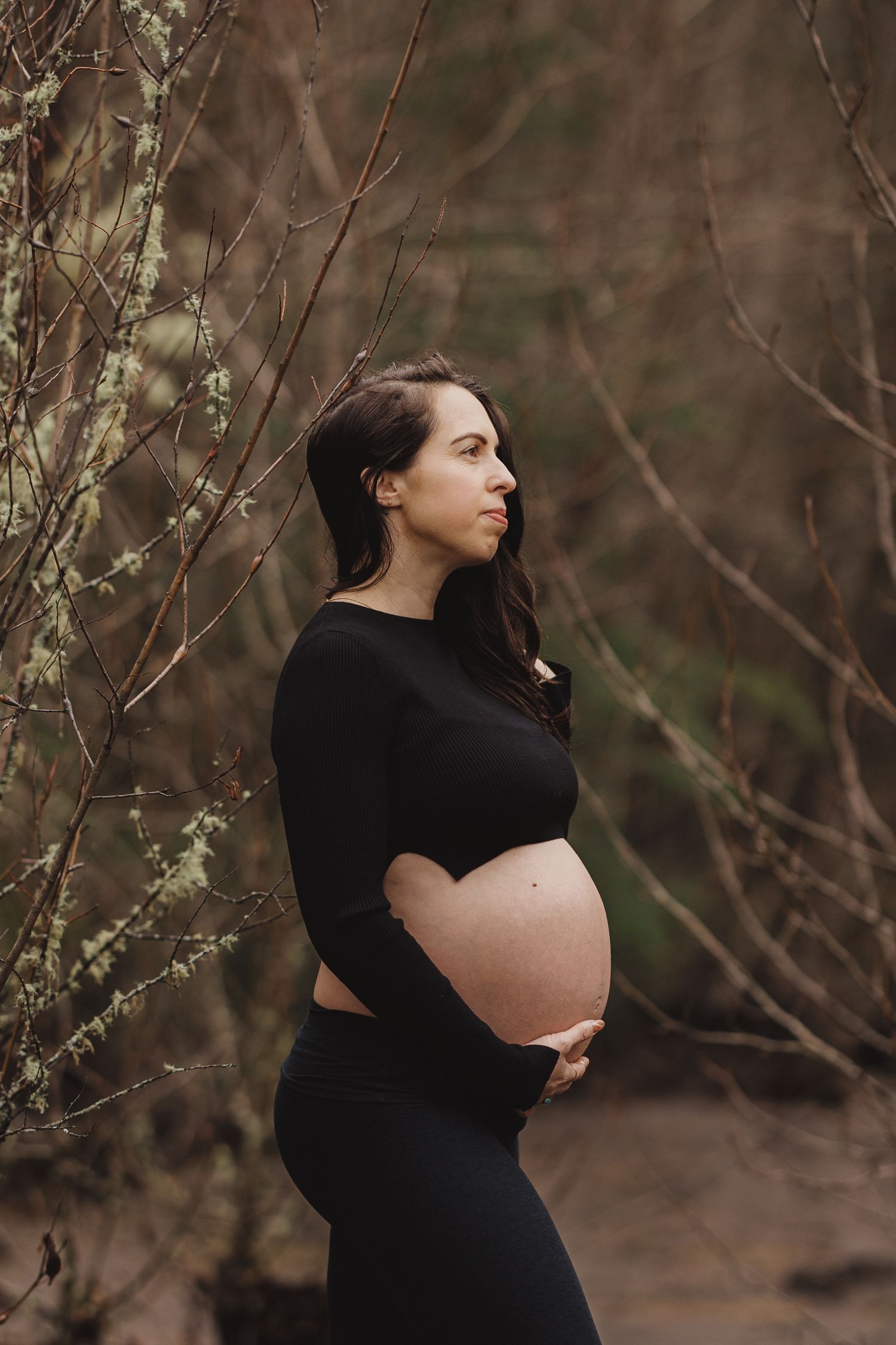 Pregnant woman in black outfit holding her bare bump while standing among bare winter branches.