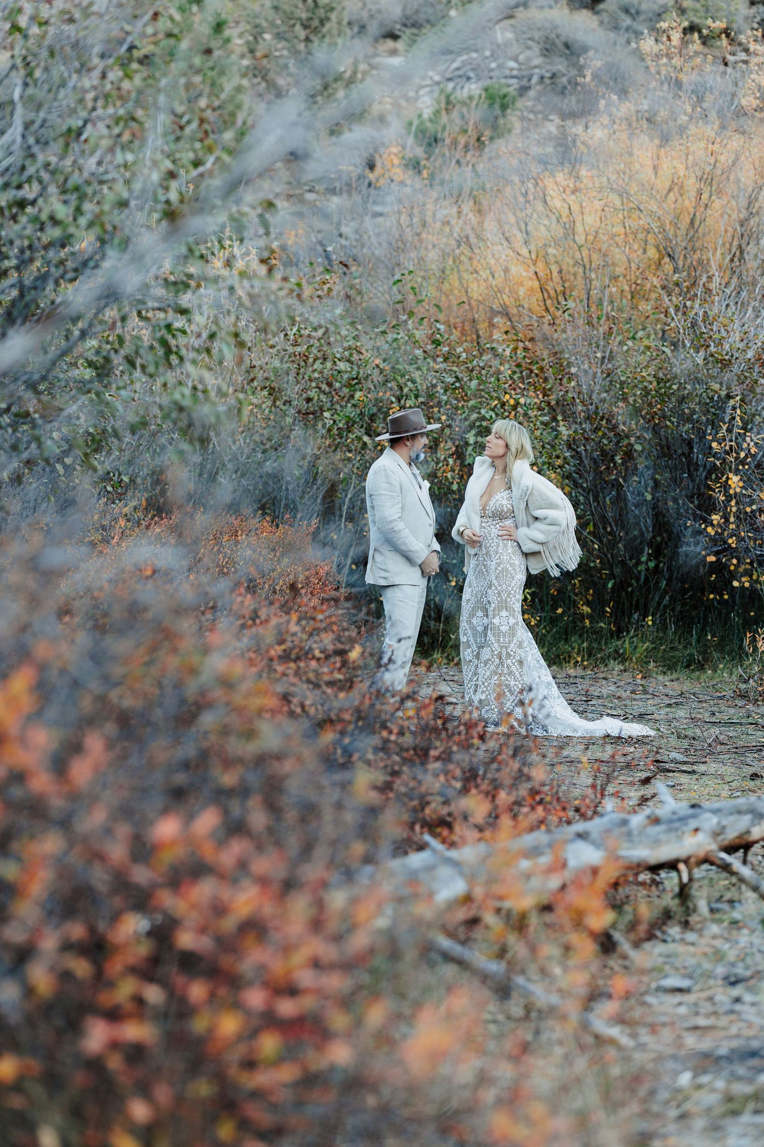 Boho couple in lace gown and tan suit walking through colorful autumn foliage landscape.