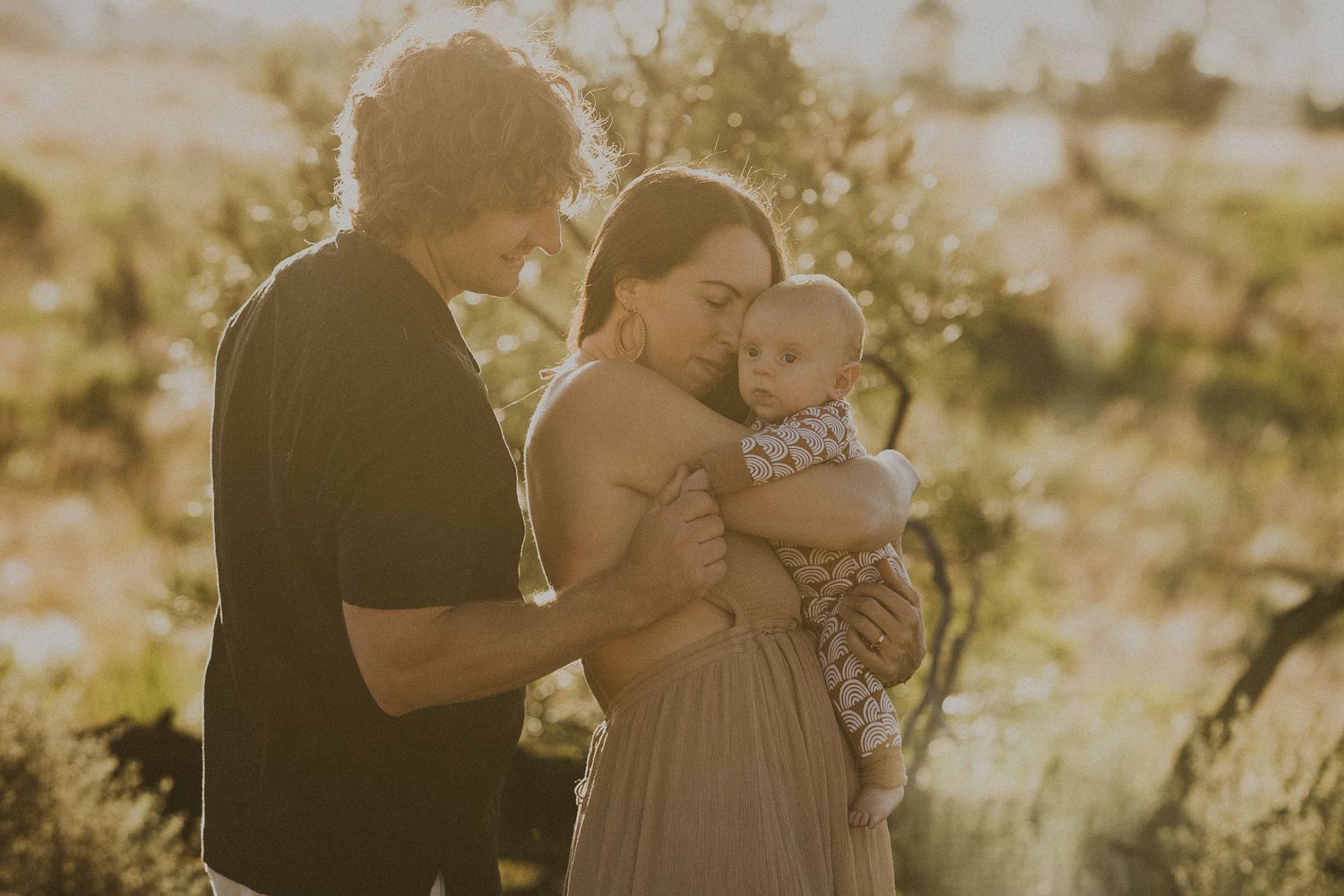 Young family with newborn baby sharing a tender moment outdoors in golden sunlight.