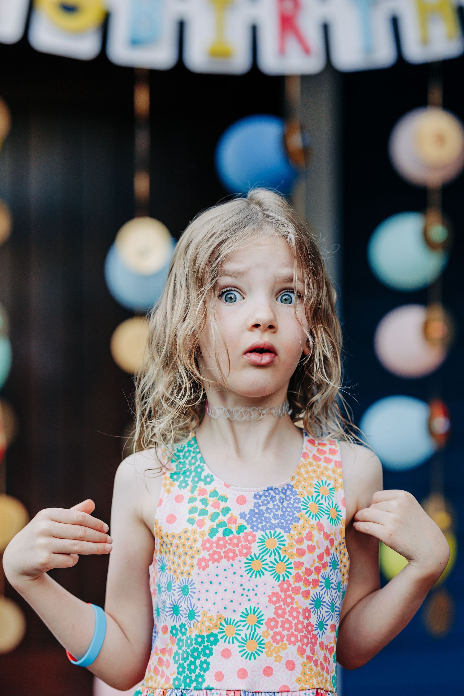 Wide-eyed young girl in colorful floral dress looks surprised at birthday party with balloon decorations.