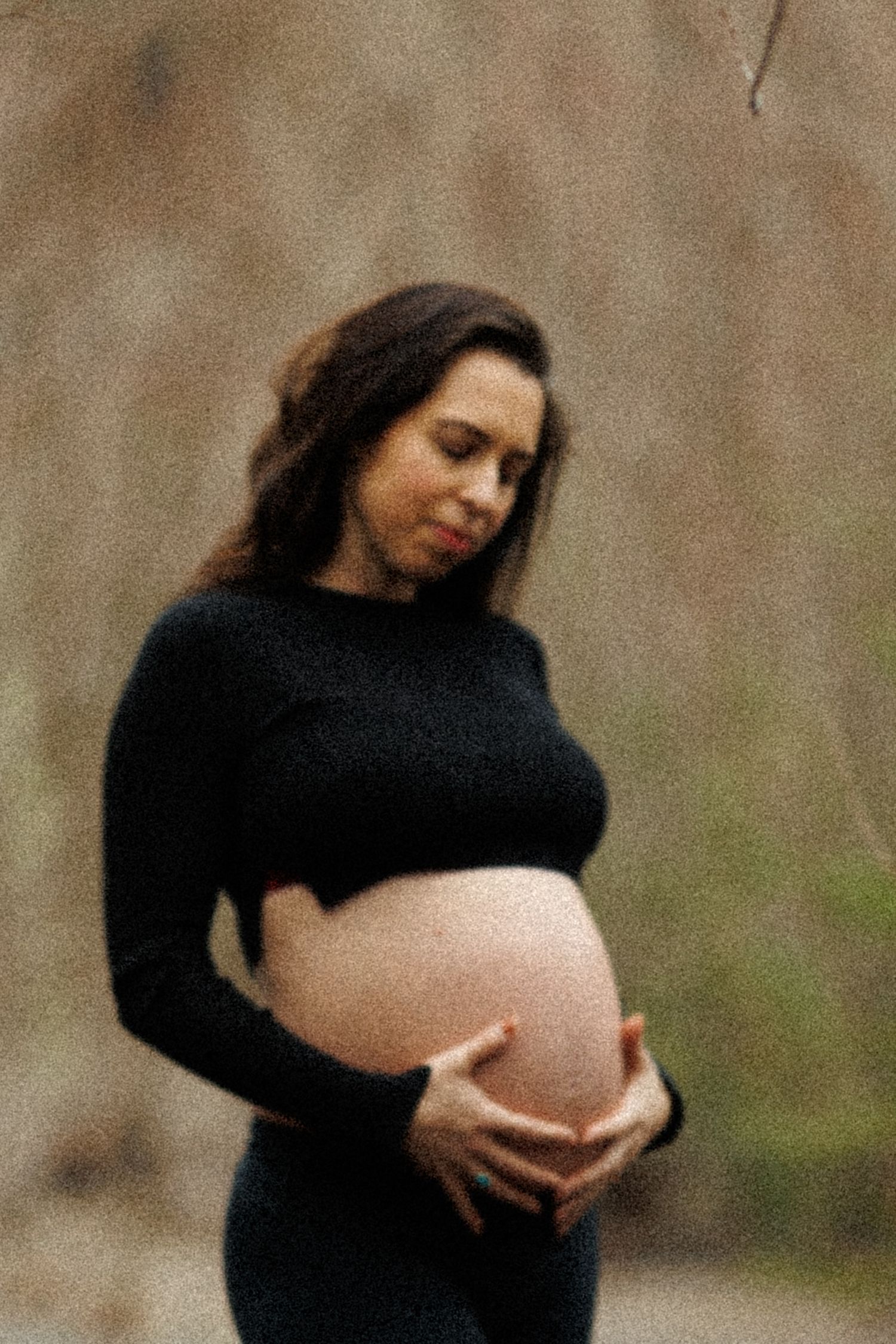Pregnant woman in black outfit holding her bare belly outdoors in a natural wooded setting.