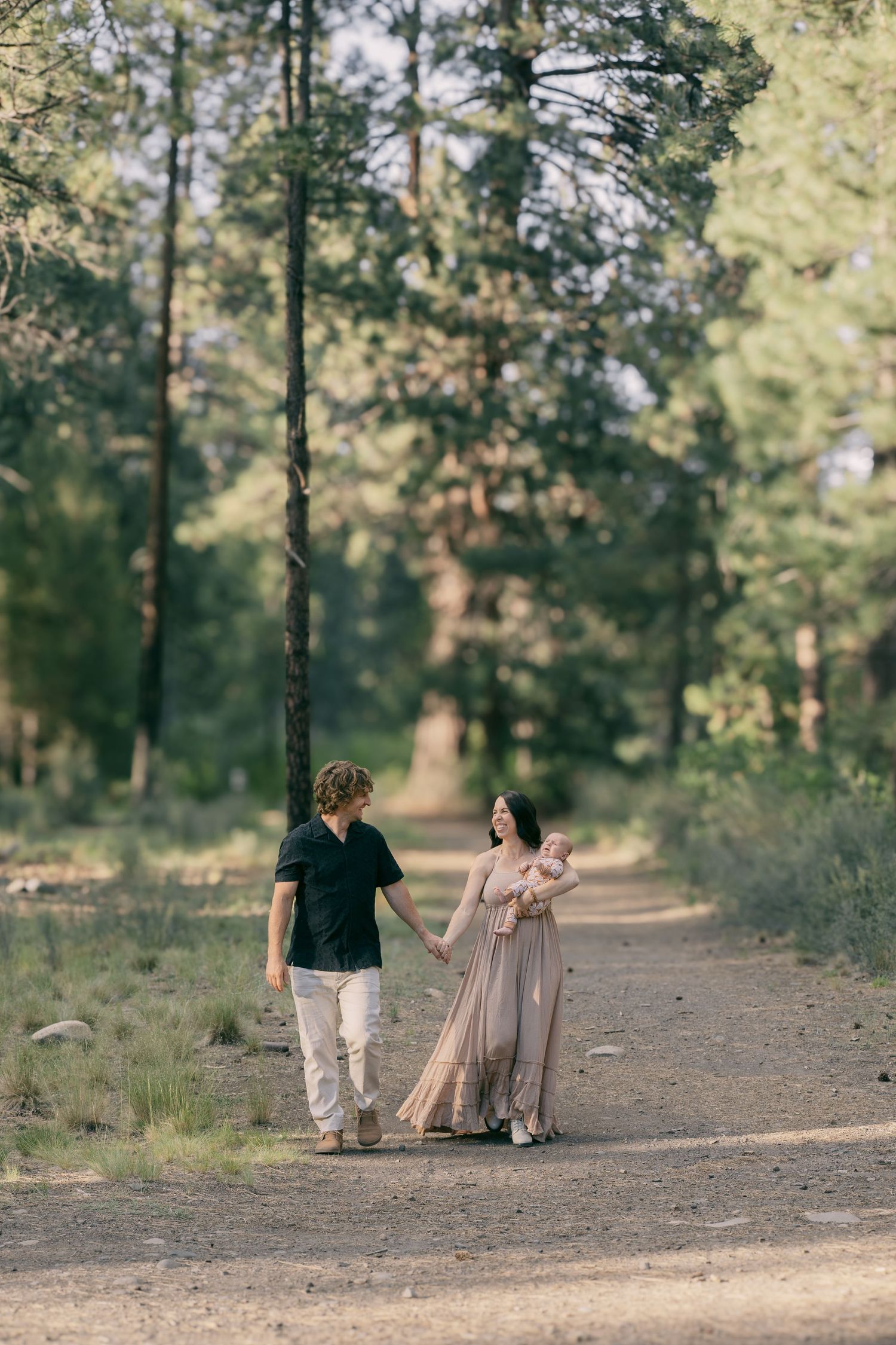 Young family of three walking together on a forest path, mother in flowing beige dress carrying newborn baby.