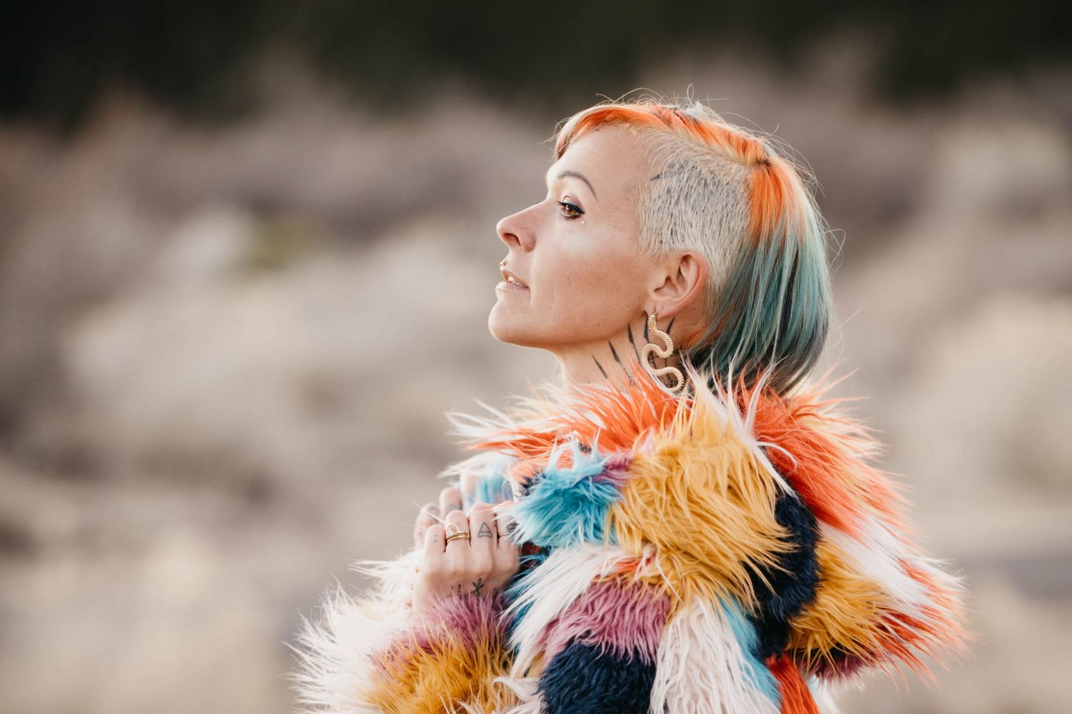 Side profile of tattooed woman in rainbow faux fur coat with shaved colorful hair.