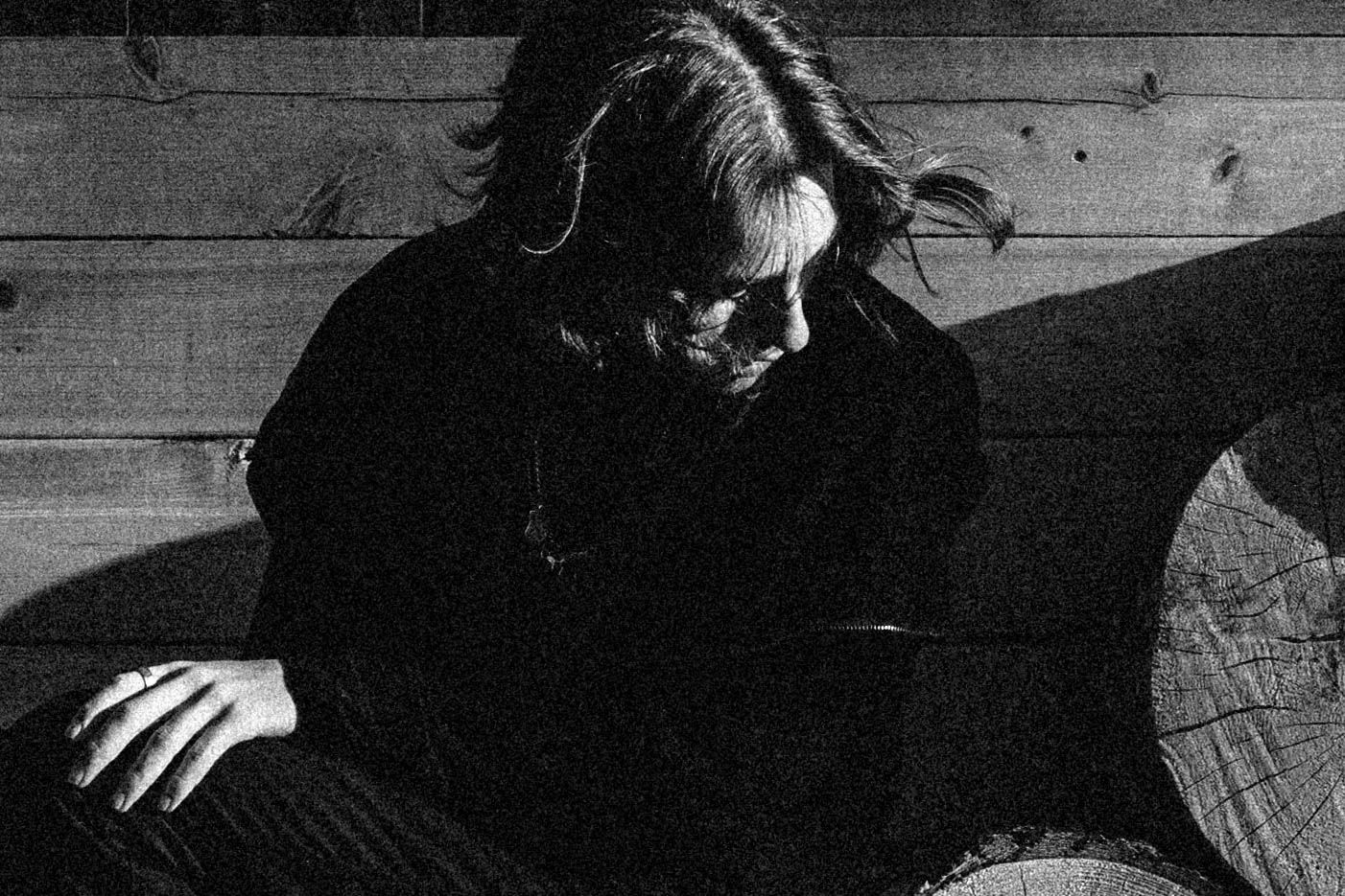 Moody black and white photo of a person with long hair sitting against a wooden wall outdoors.