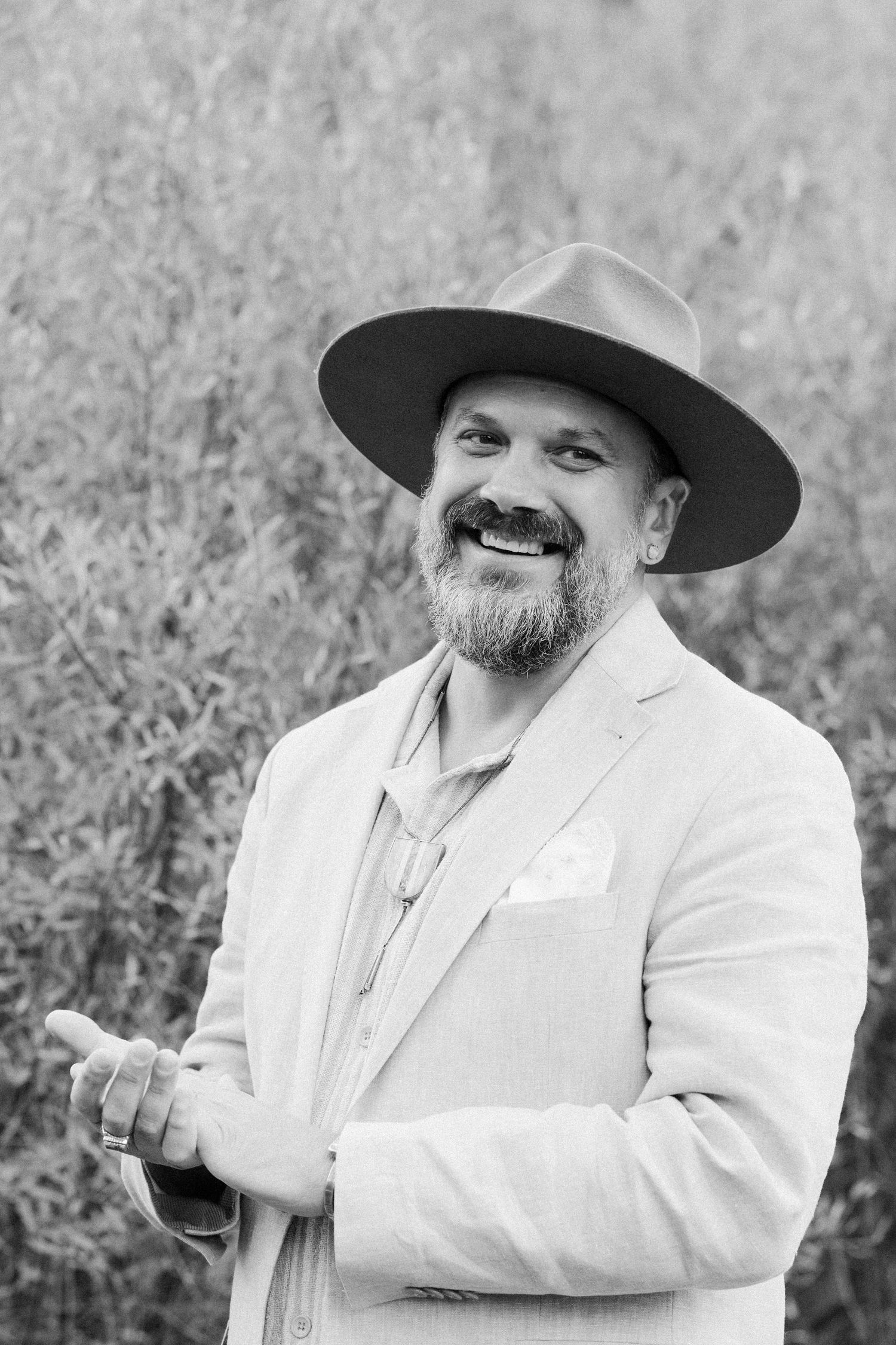 Smiling bearded man in wide-brim hat and linen blazer posing outdoors in black and white photo.