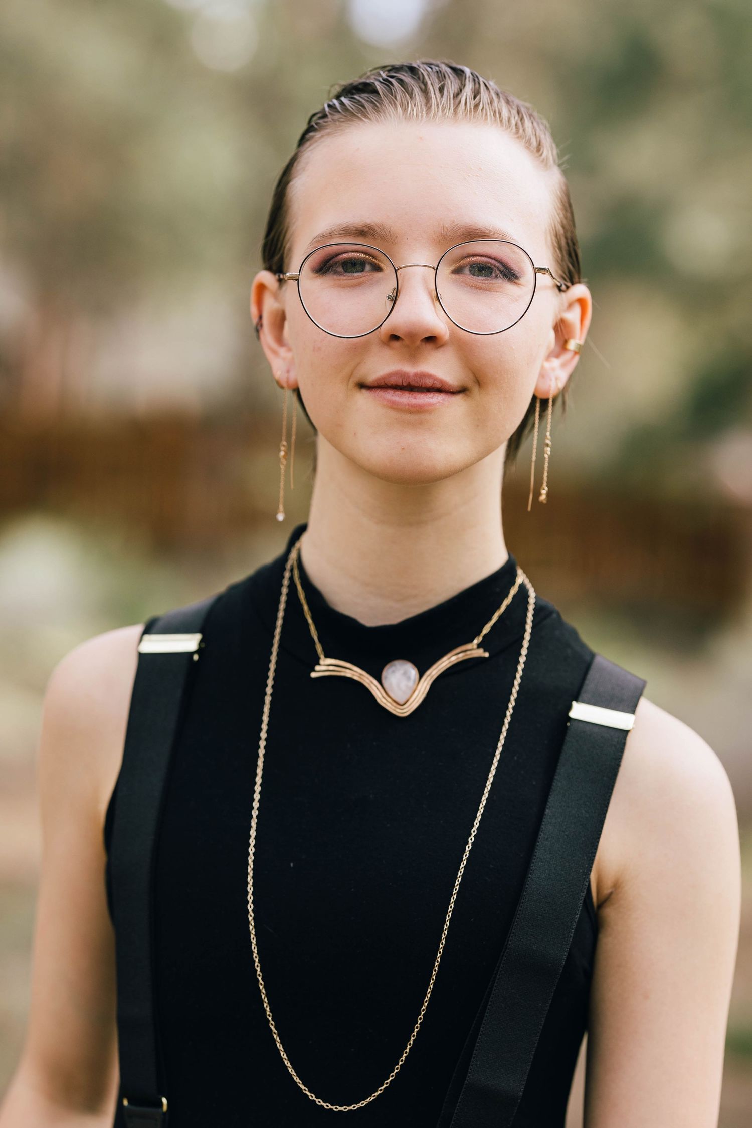 Young person smiling in round glasses wearing black sleeveless top with elegant gold layered necklaces outdoors.