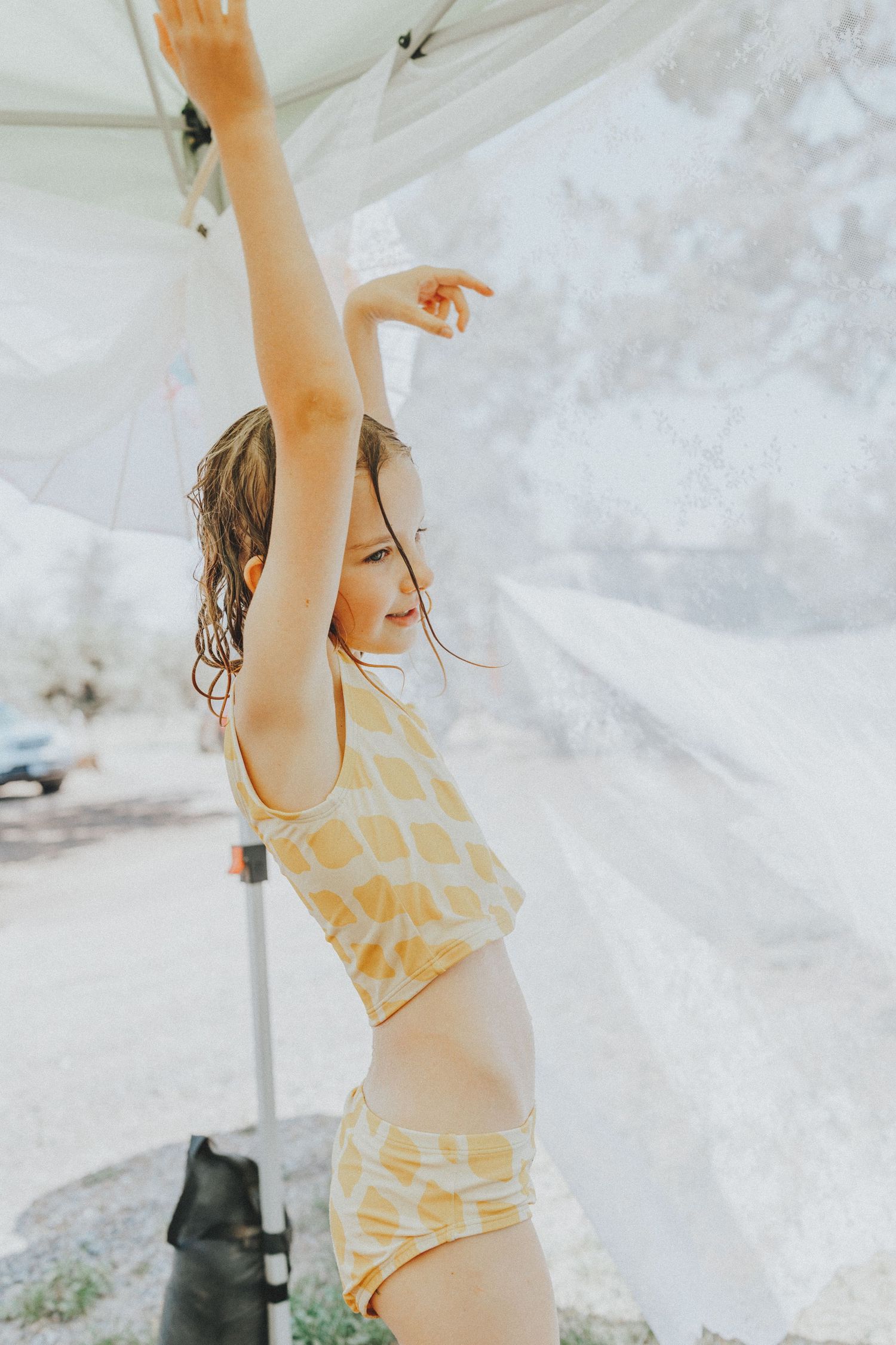 A young girl in a yellow patterned bikini reaches up toward a white canopy outdoors.