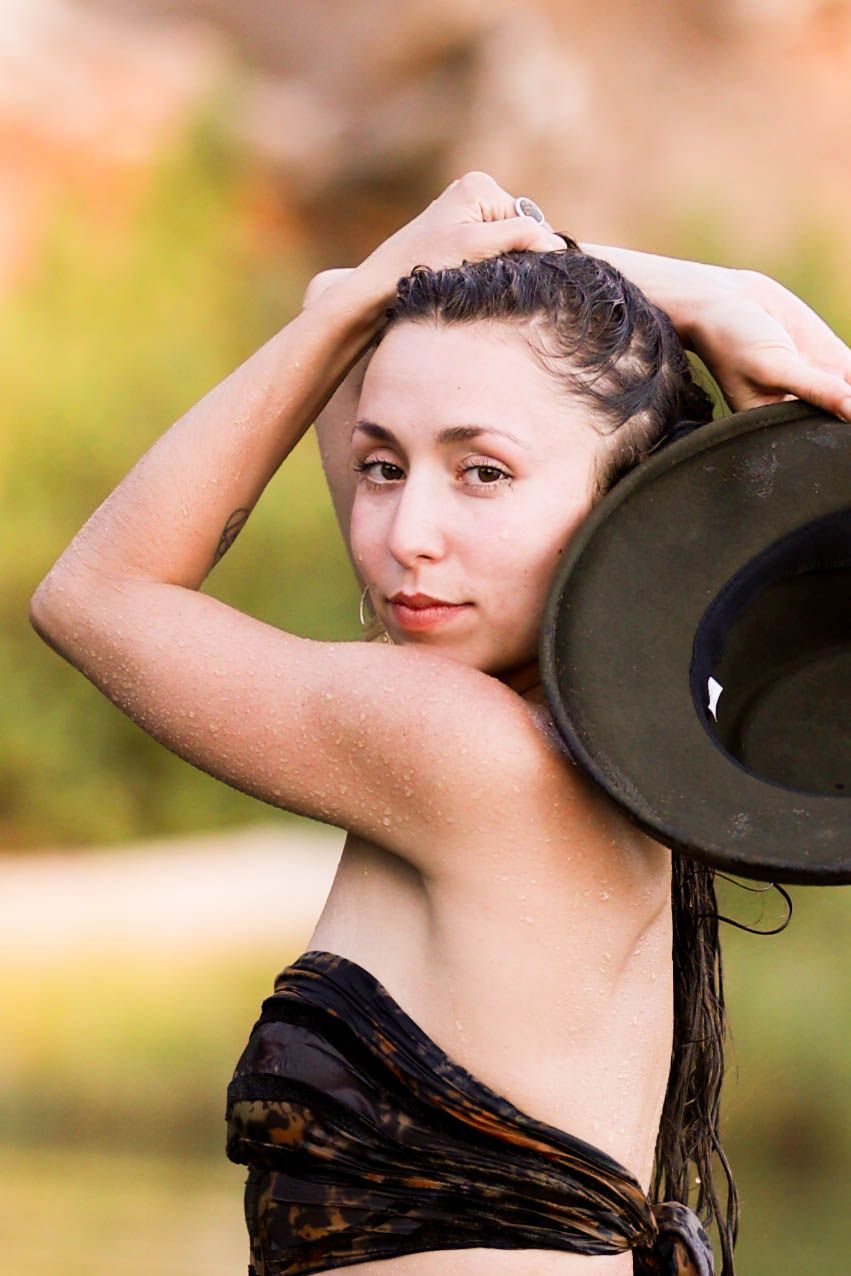 Woman in animal print bikini top holds green hat above head with wet hair in natural outdoor setting.