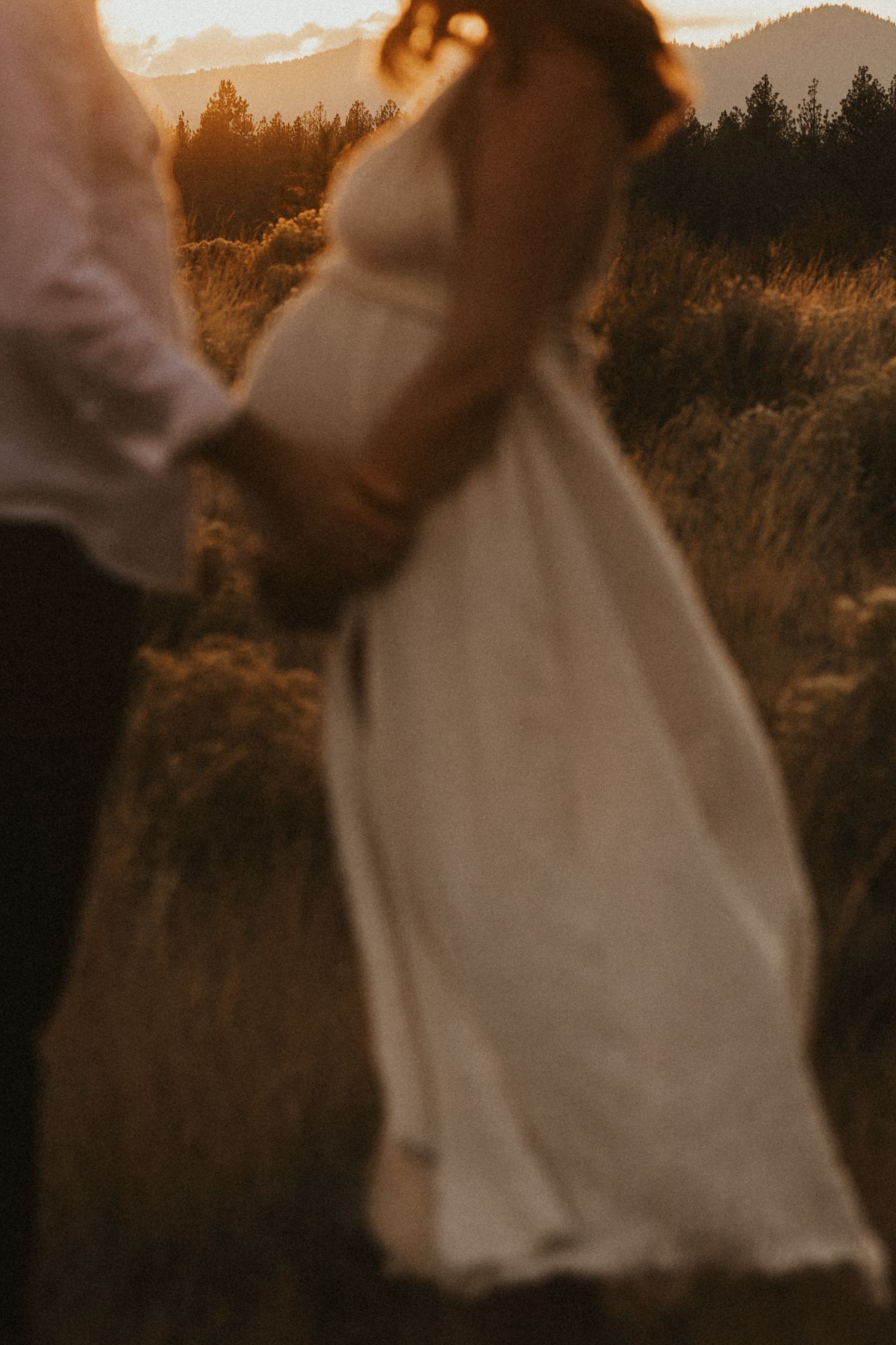 Blurred artistic maternity photo of couple holding hands in glowing sunset field setting.