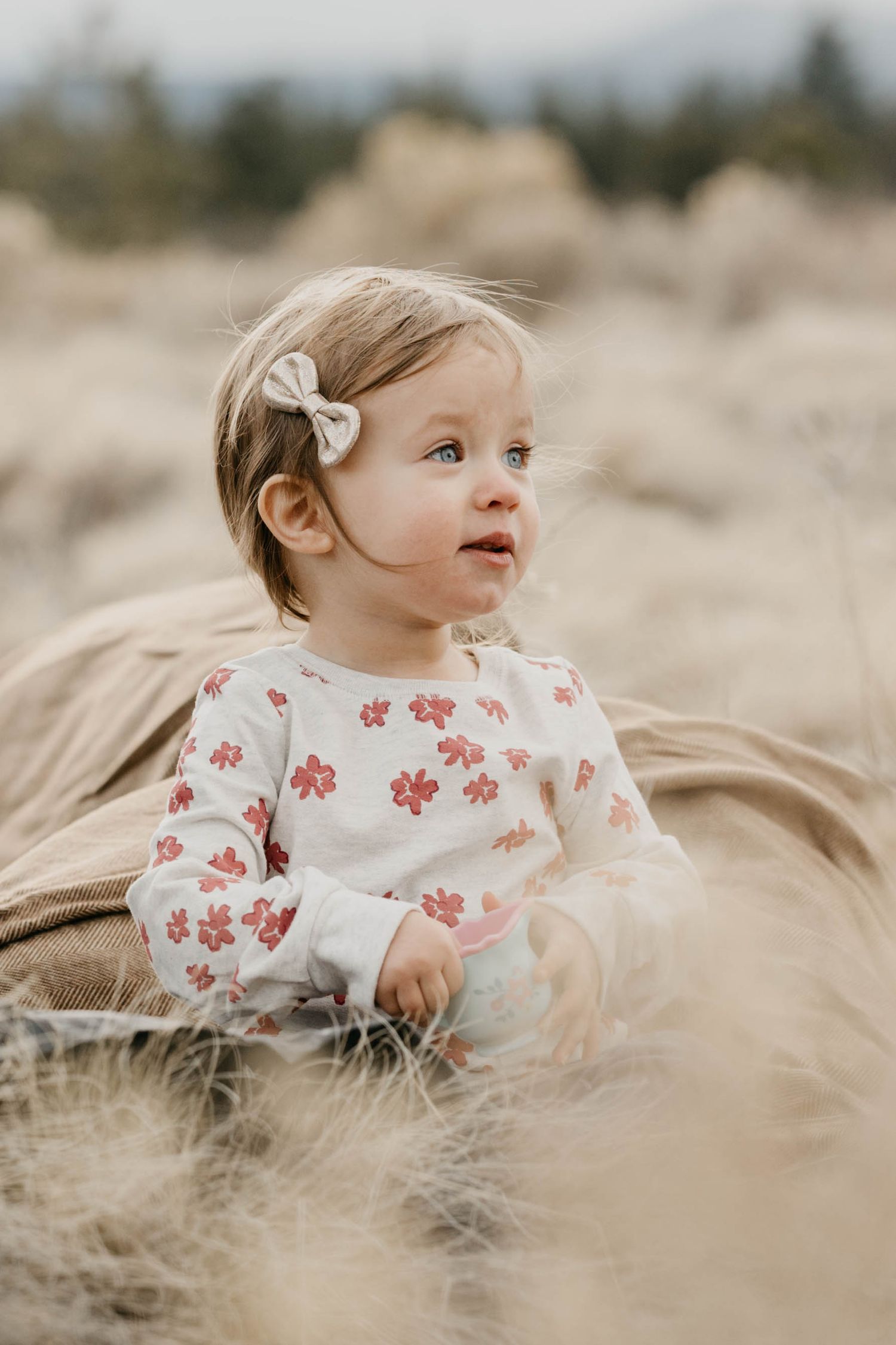 Sweet baby girl in floral outfit sitting on a blanket in a golden field.