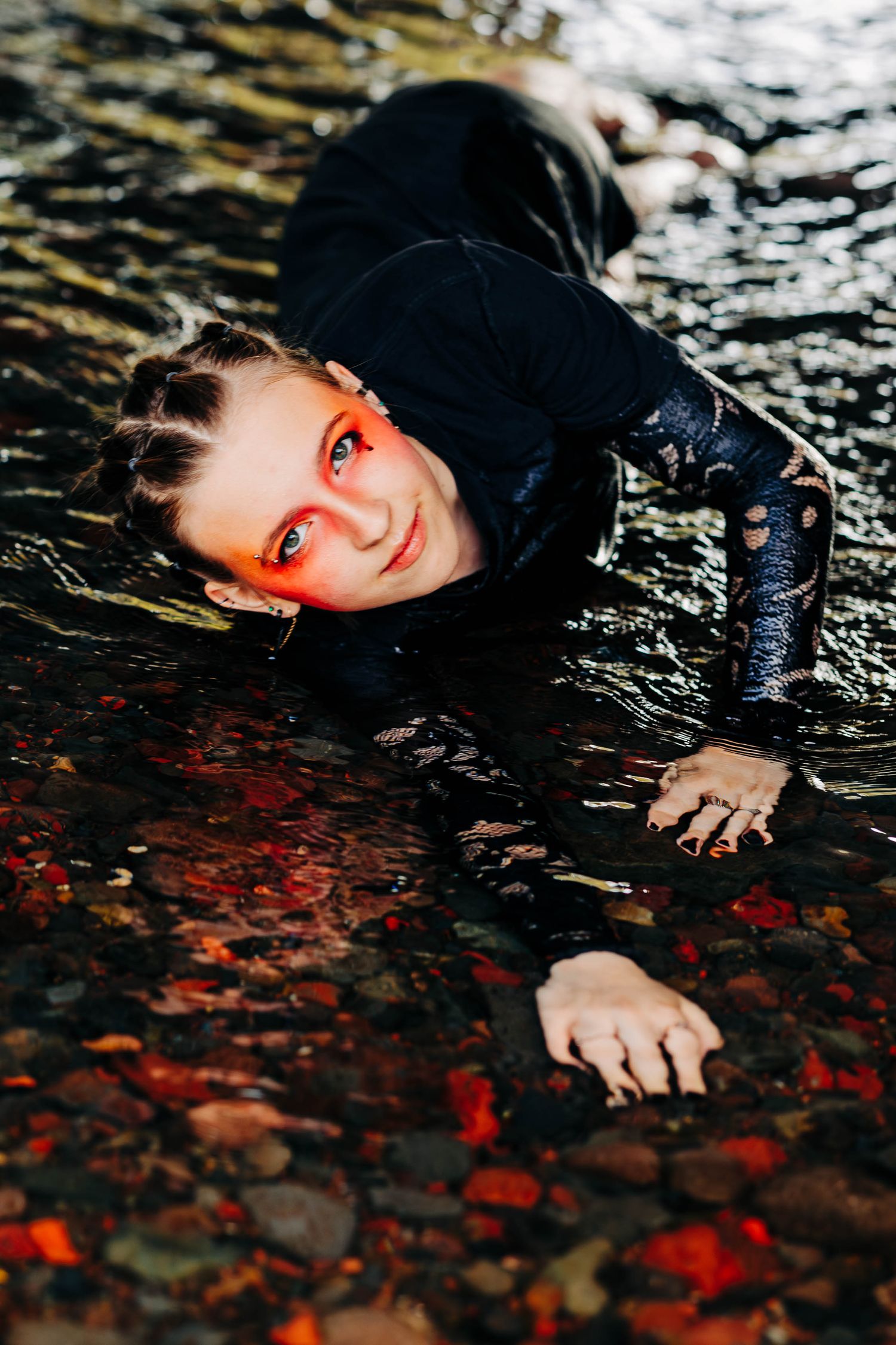 Woman with dramatic red makeup crawls through shallow water in black lace outfit.