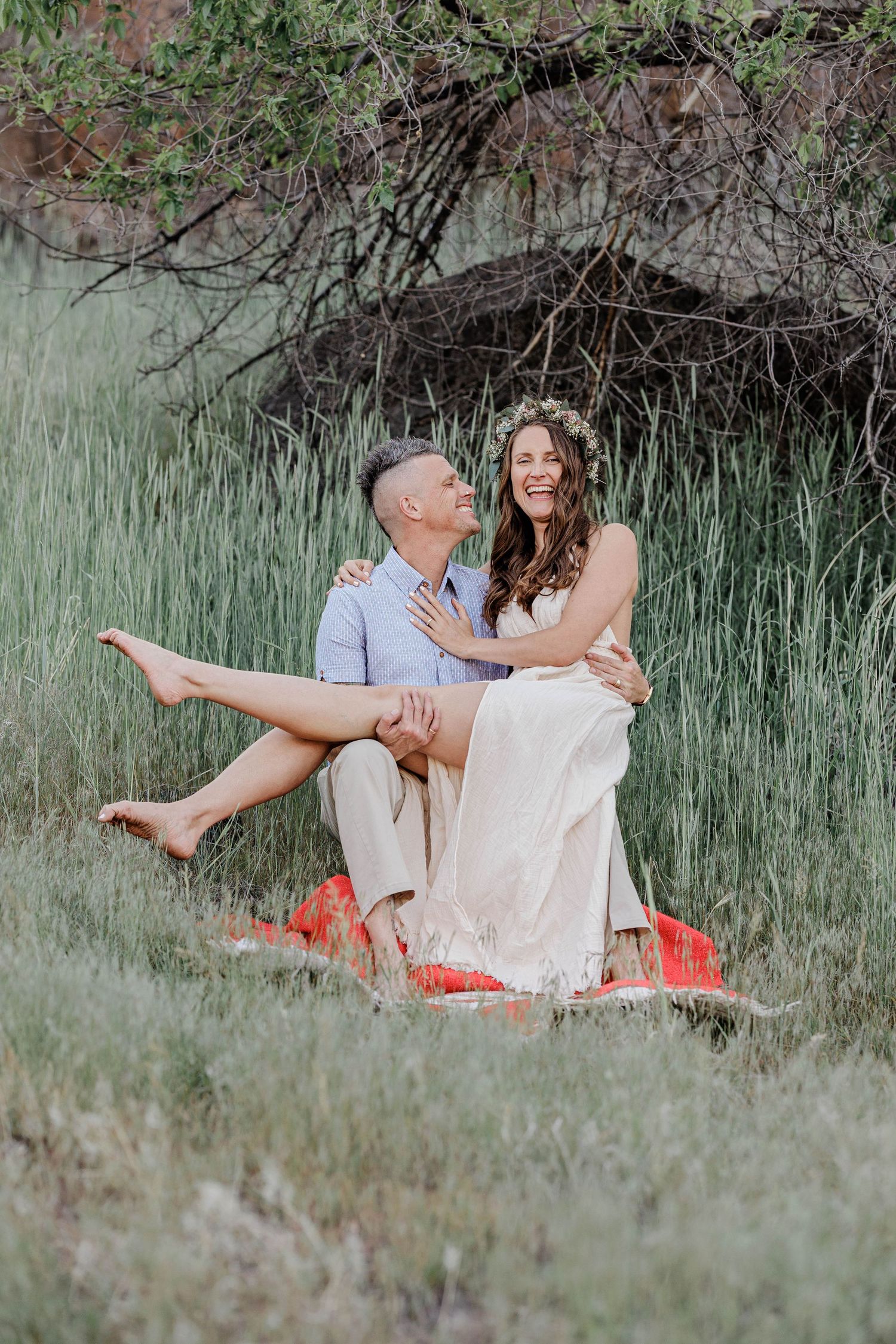 Groom holds laughing bride in floral crown on red blanket in lush green grass.