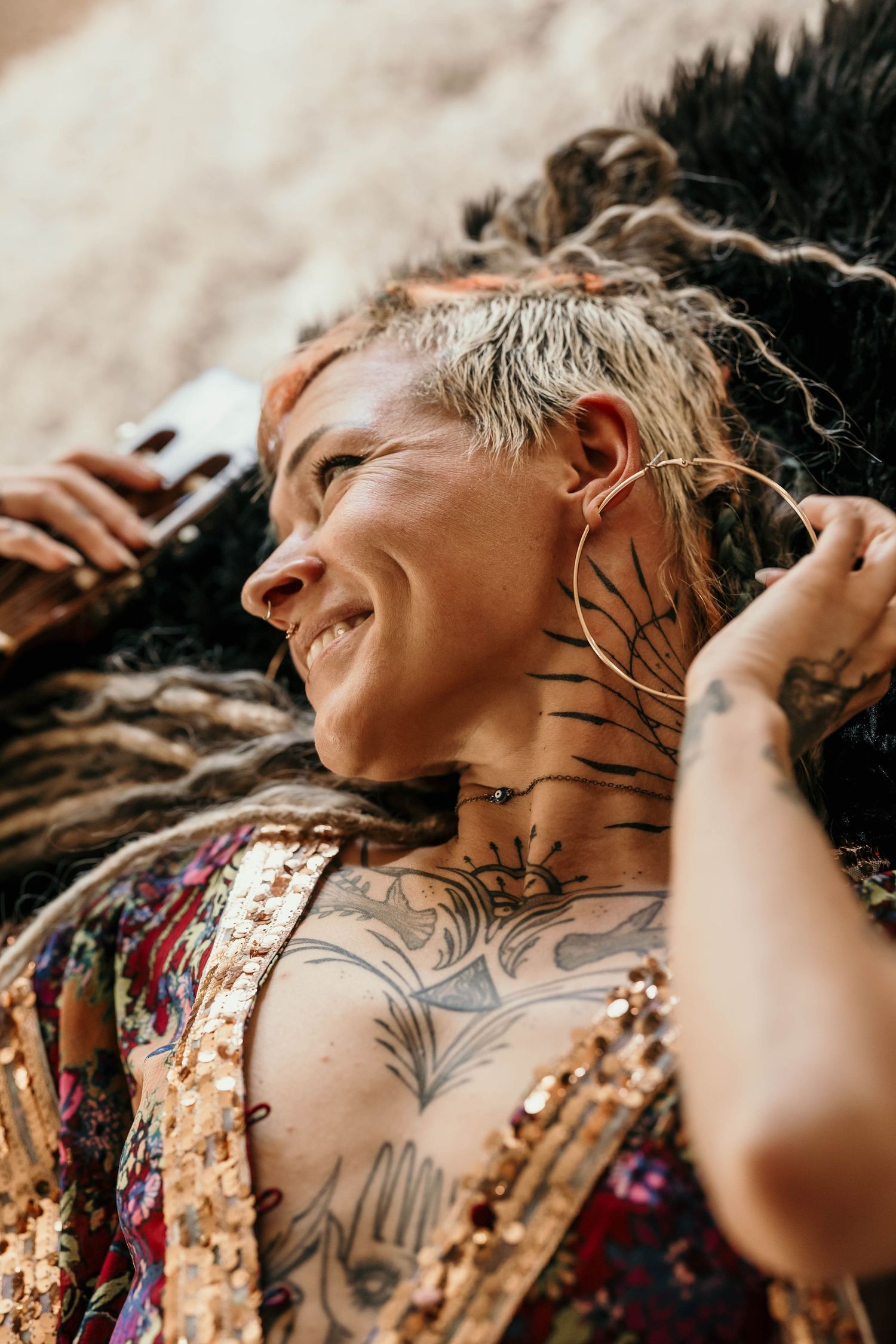 Smiling tattooed woman with dreadlocks and neck tattoos lying outdoors wearing a sequined top.