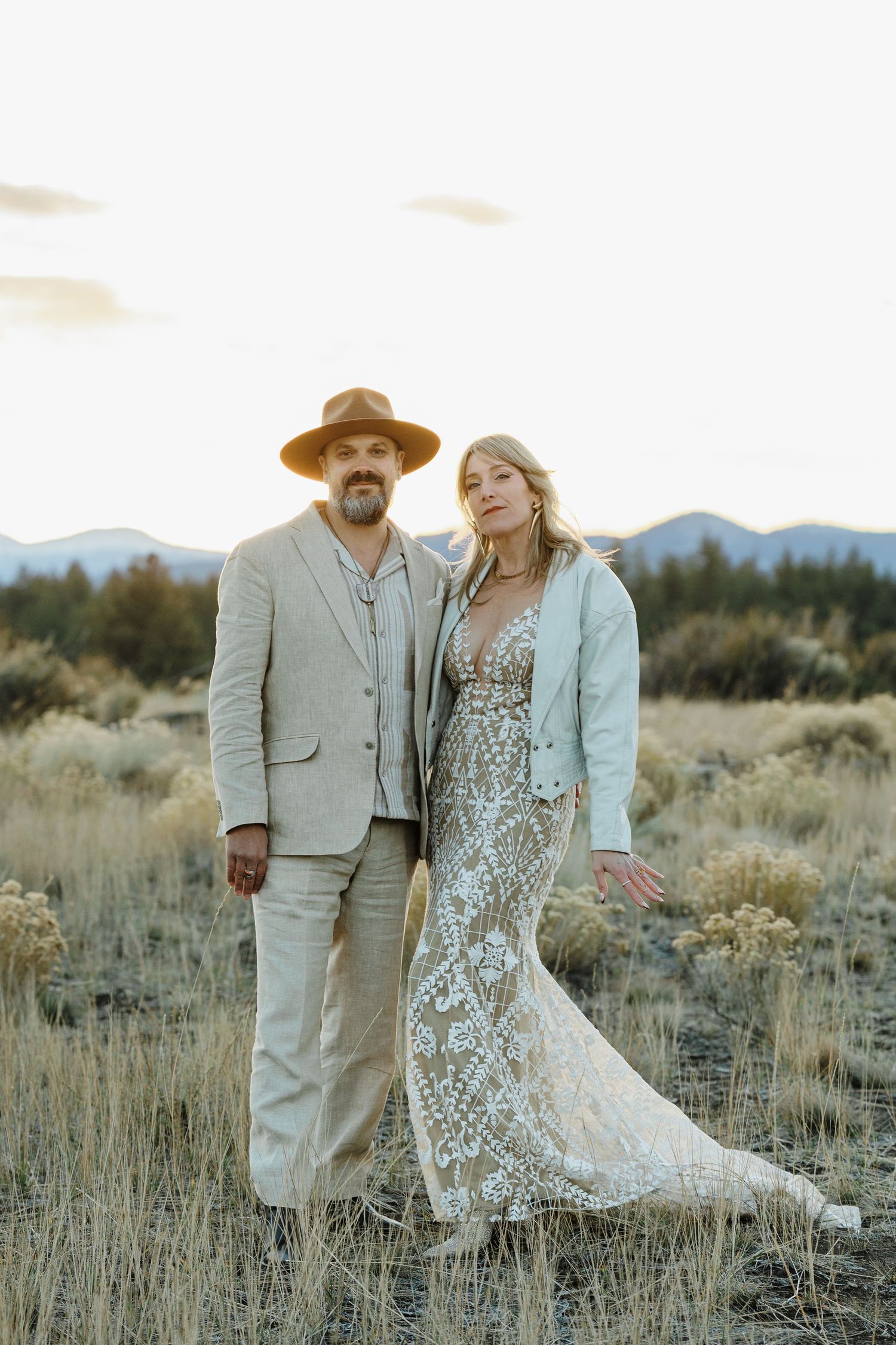 Couple in boho wedding attire poses in golden field with mountain backdrop at sunset.