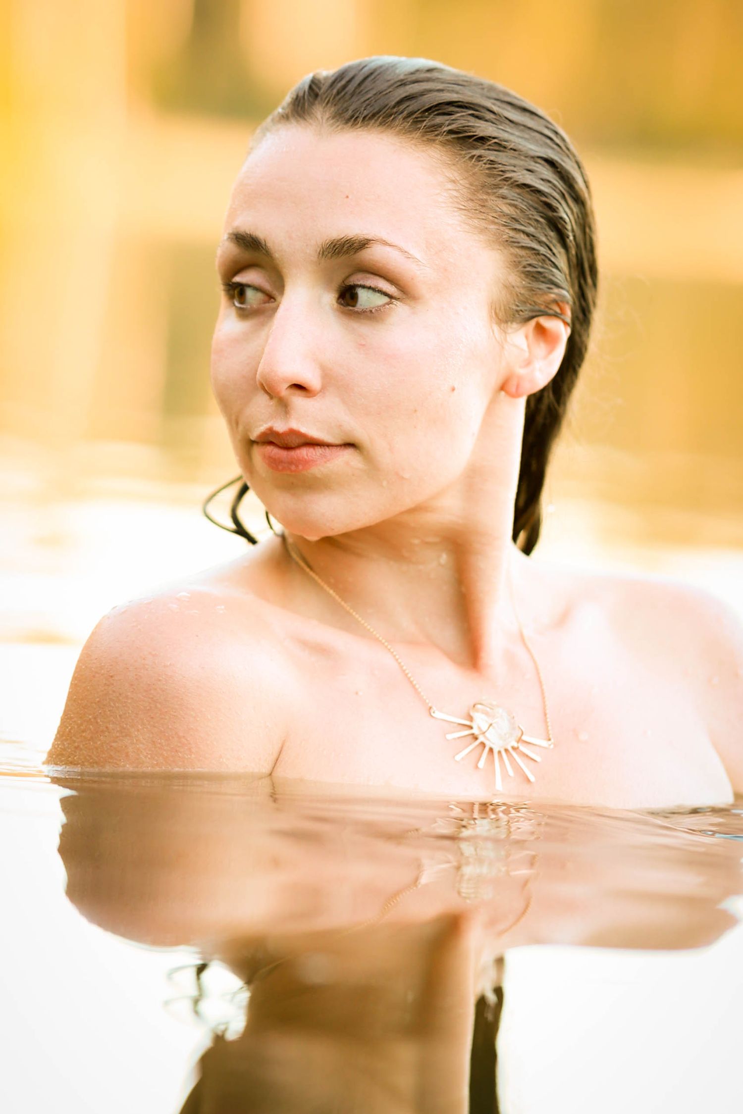 Woman with slicked back wet hair emerges from golden reflective water wearing a delicate sun pendant necklace.