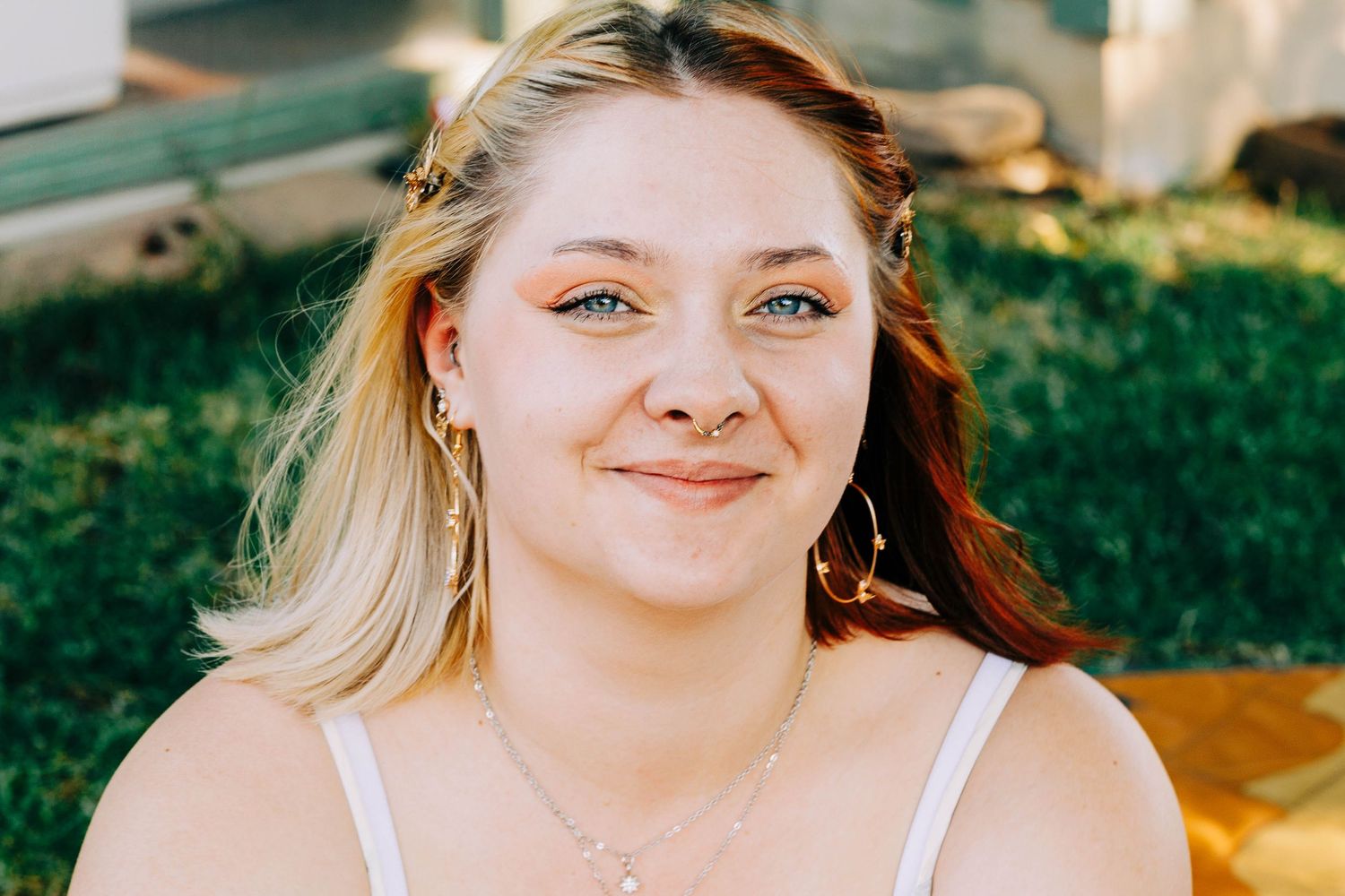 Young blonde woman with blue eyes smiling outdoors wearing a white top and delicate chain necklace.