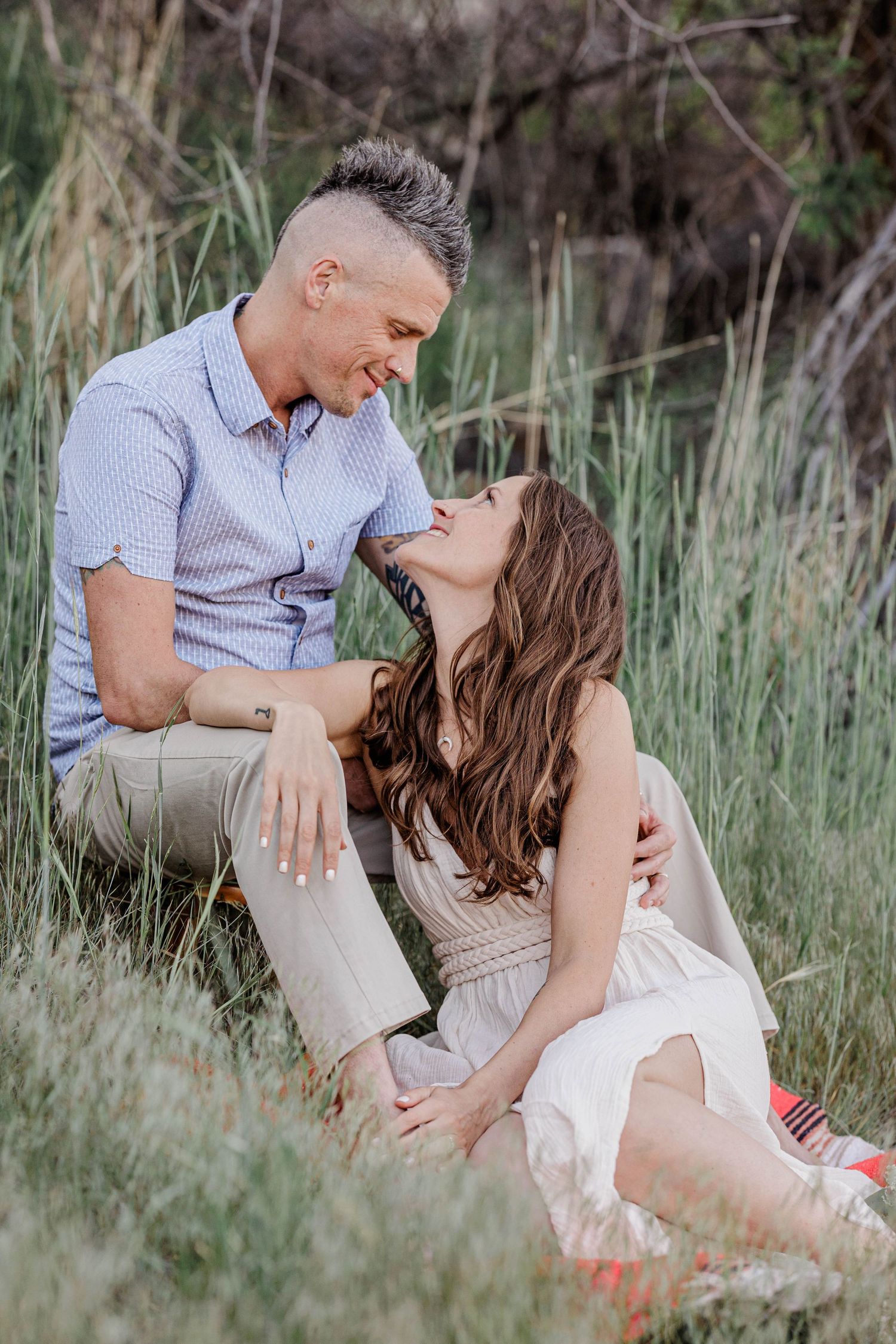Couple gazes lovingly at each other while sitting together in tall green grass.