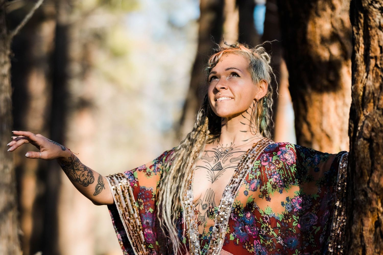 Tattooed bohemian woman with long dreadlocks smiling in a sunlit forest wearing a floral kimono.