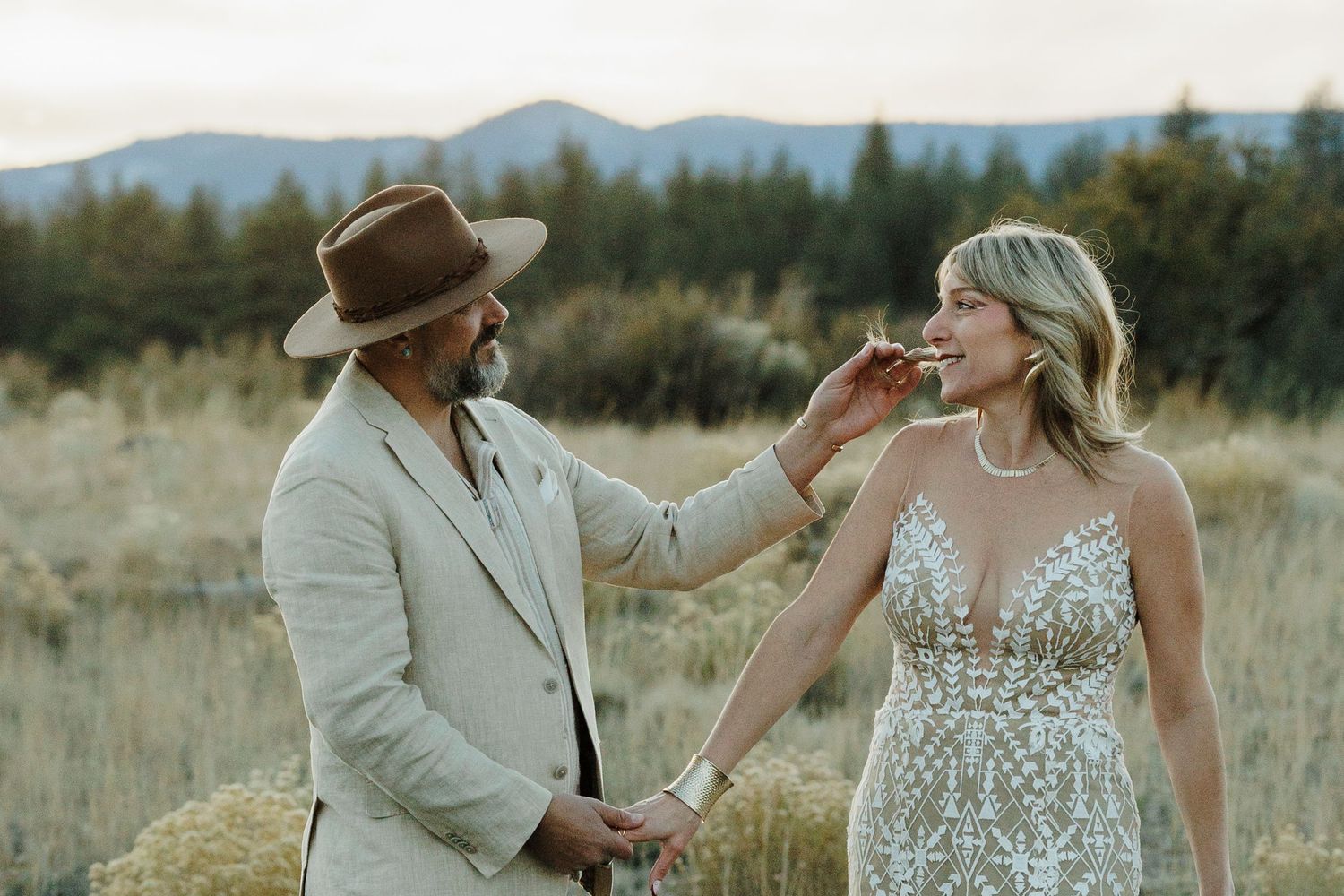Groom in tan suit and hat feeds bride in lace wedding dress in a scenic field.