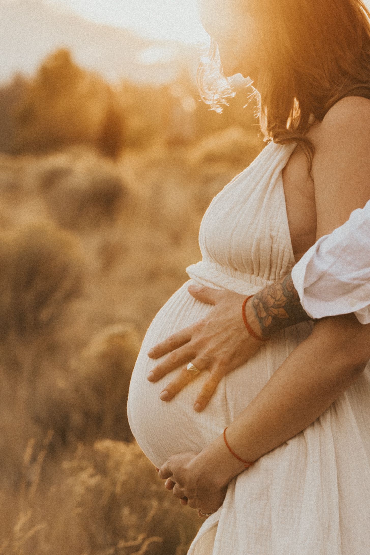 Close-up of pregnant woman's belly with tattooed hands cradling bump during golden sunset shoot.