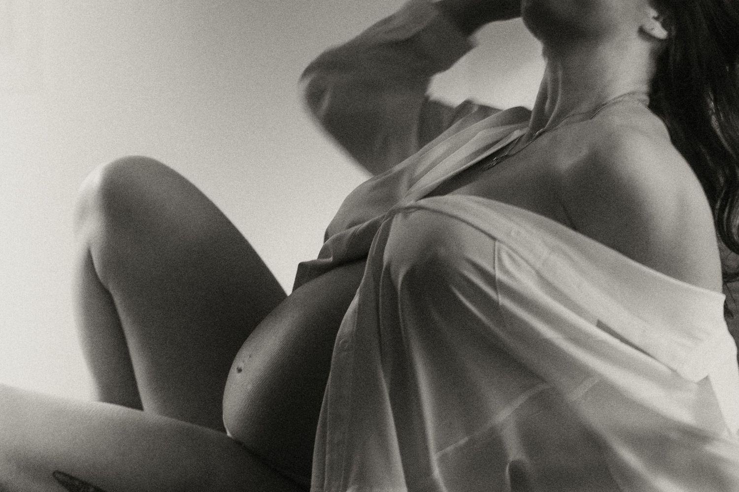 Black and white photo of pregnant woman in sheer wrap sitting and gazing upward showing bare bump.