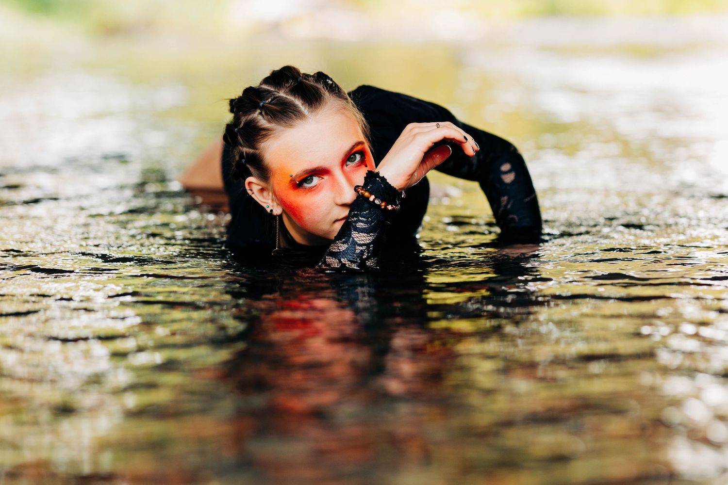 Woman with striking red makeup poses partially submerged in water with braided hair.