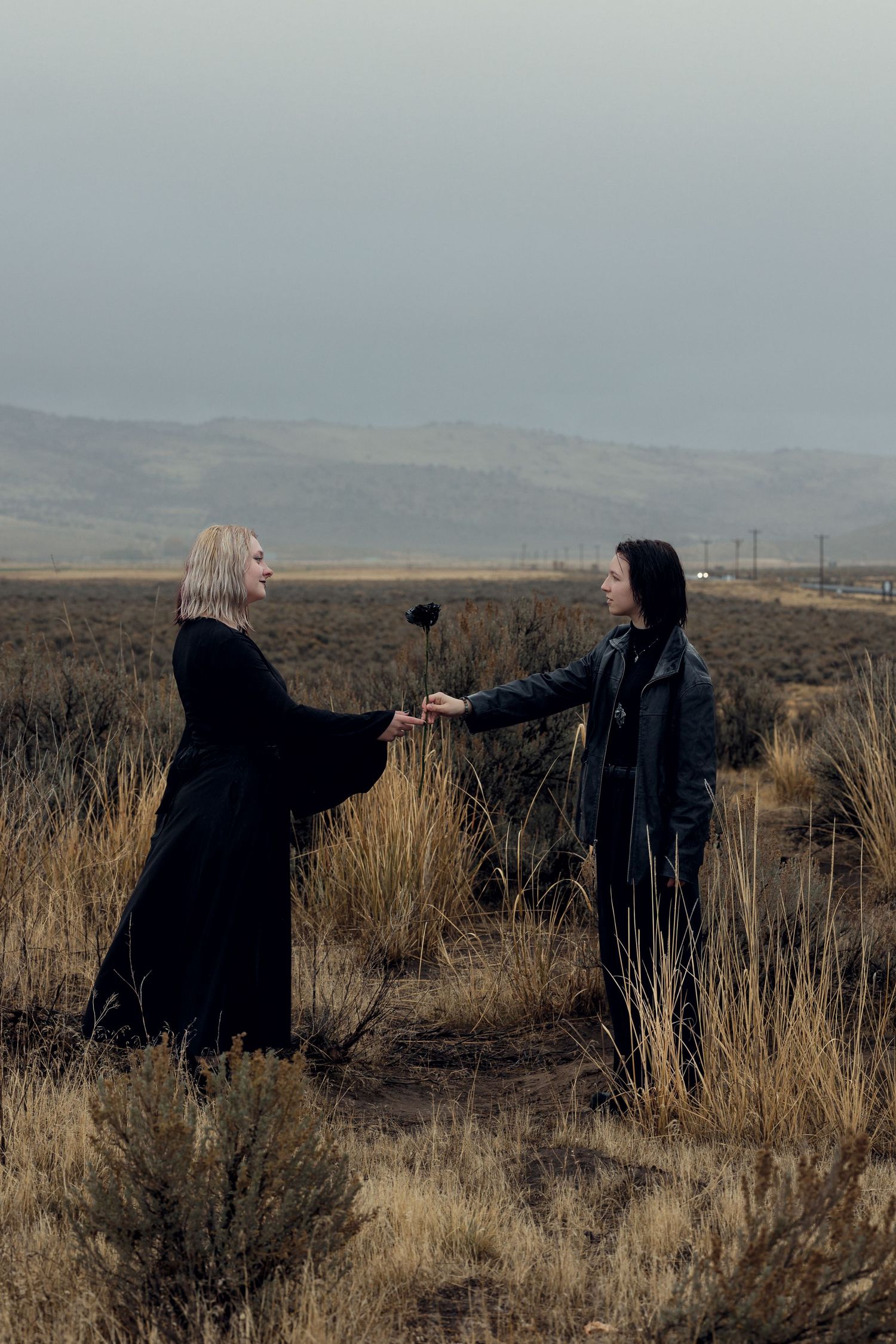Two women in black clothing exchange a dark flower in a misty open field with mountains in the background.