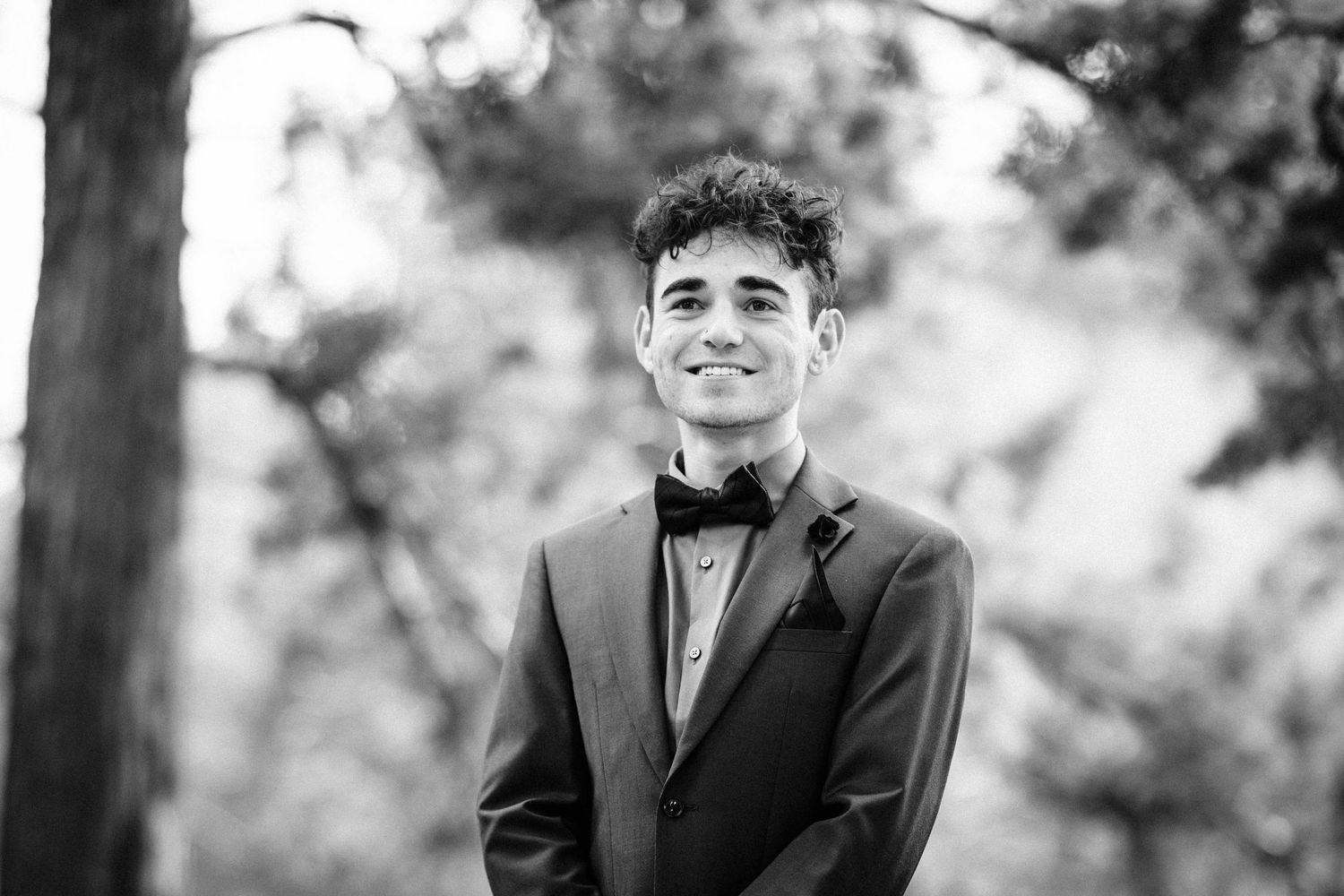 Black and white portrait of smiling young man in suit and bow tie standing outdoors near trees.