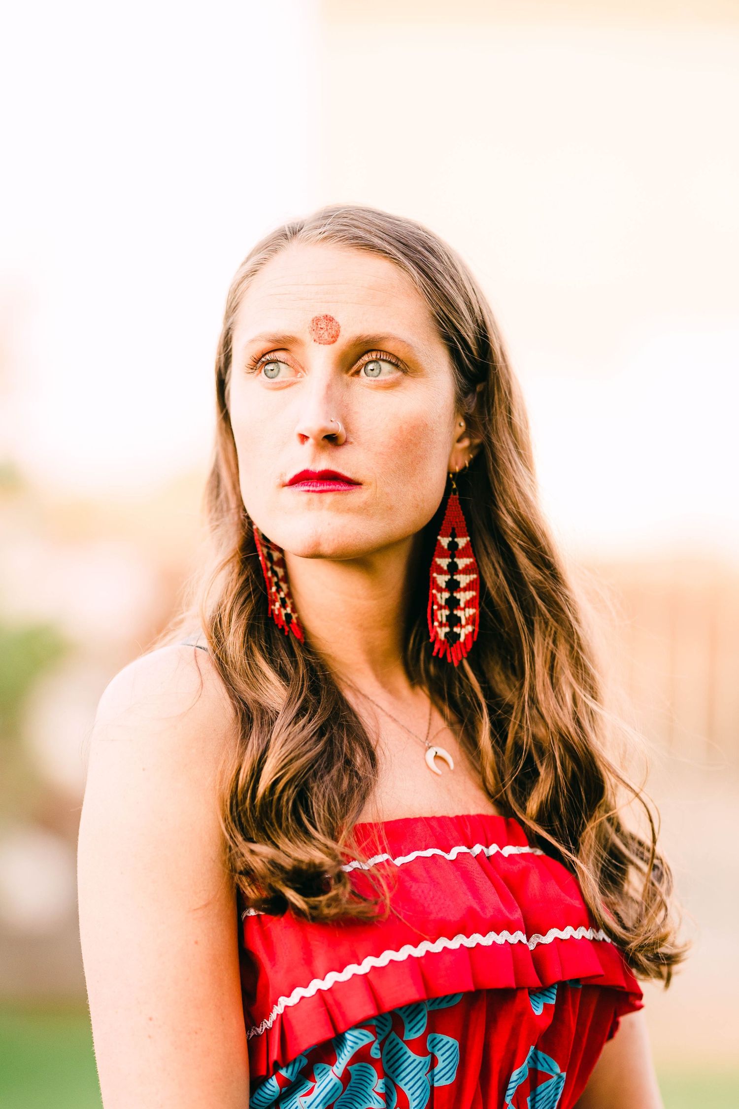 Woman in red off-shoulder dress with decorative earrings and forehead marking gazing upward outdoors.