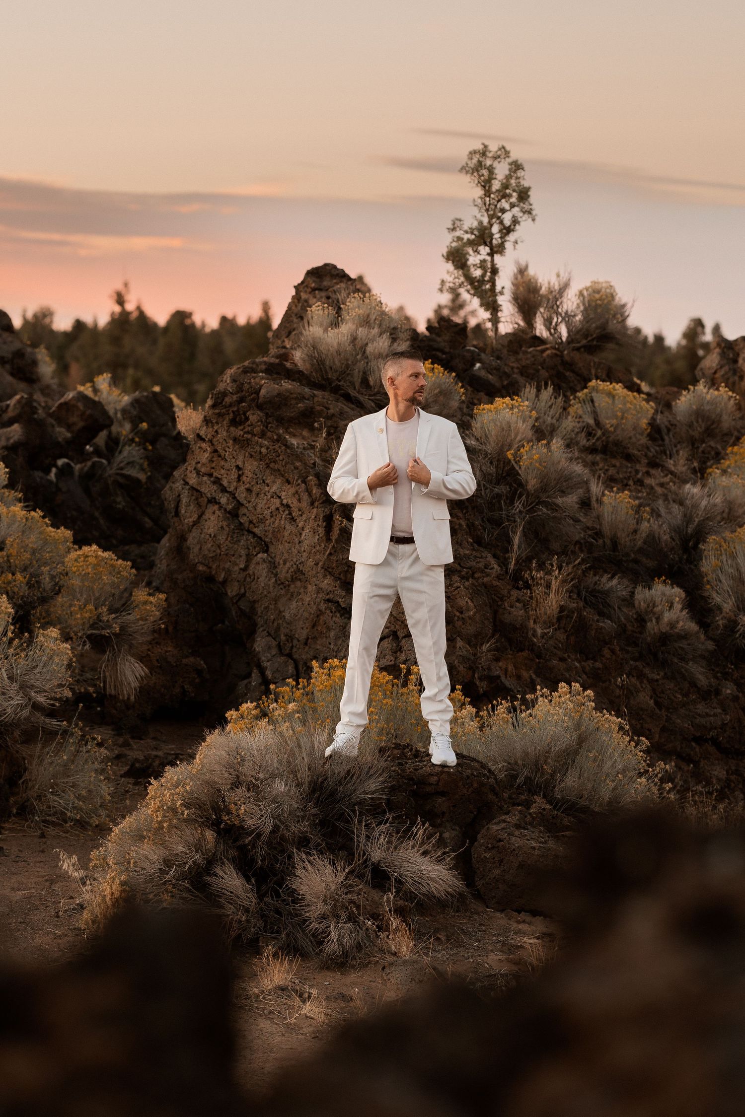 Man in white suit standing on rocky terrain with sagebrush at pink sunset.