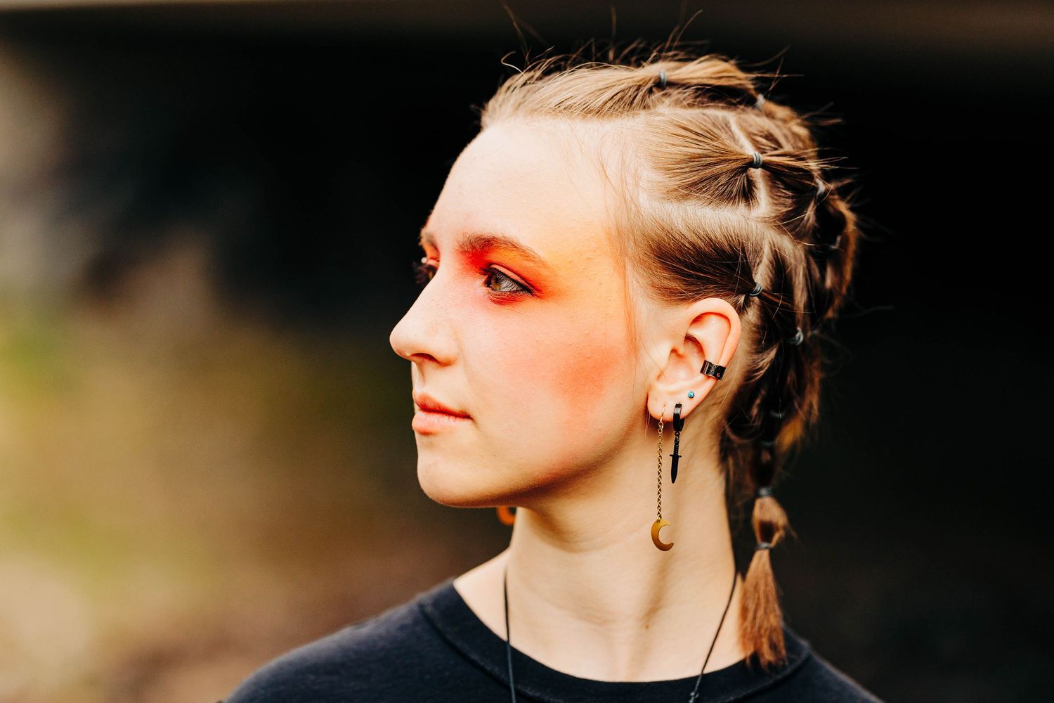 Young woman with braided blonde hair and bold orange-red eye makeup, wearing multiple ear piercings with feather earrings.