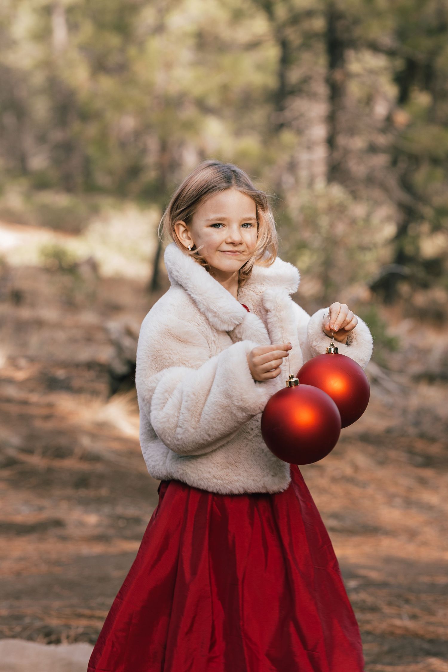 Smiling girl in white fluffy jacket and red skirt holding large red Christmas ornament balls in forest.