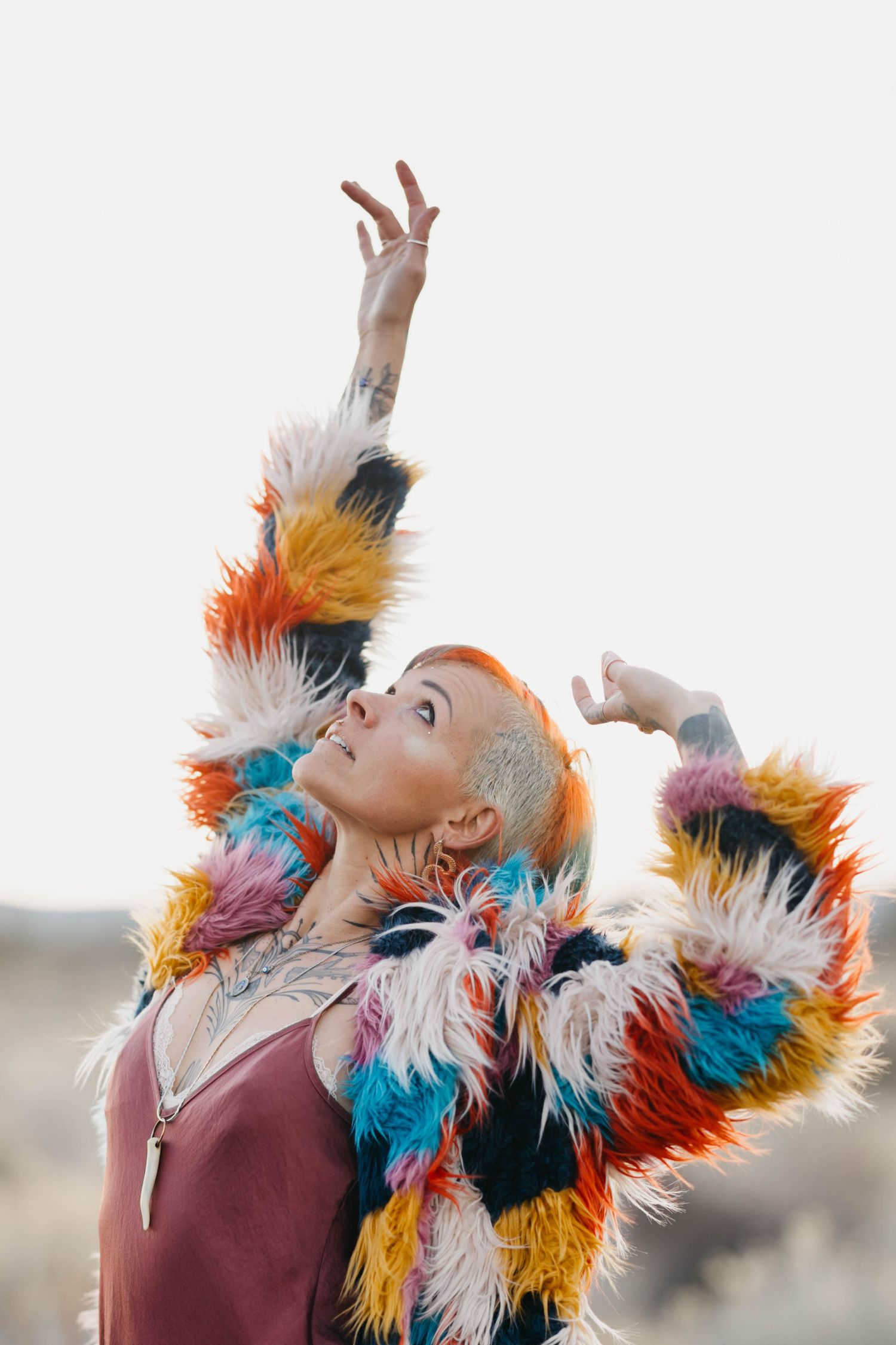 Tattooed woman in colorful faux fur coat reaches arm upward against bright sky.