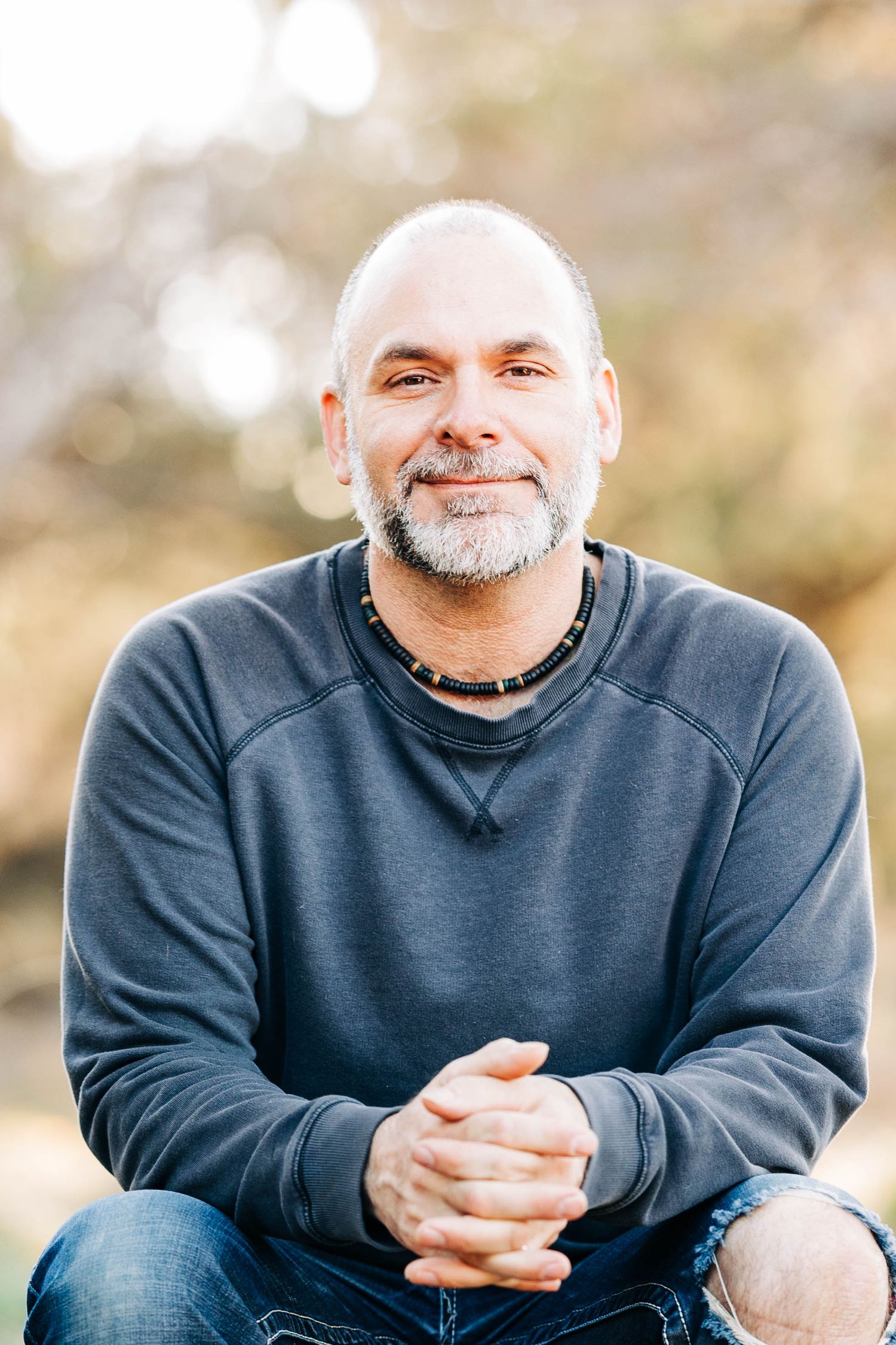 Smiling bald man with a gray beard sitting outdoors in a gray sweatshirt with hands clasped.